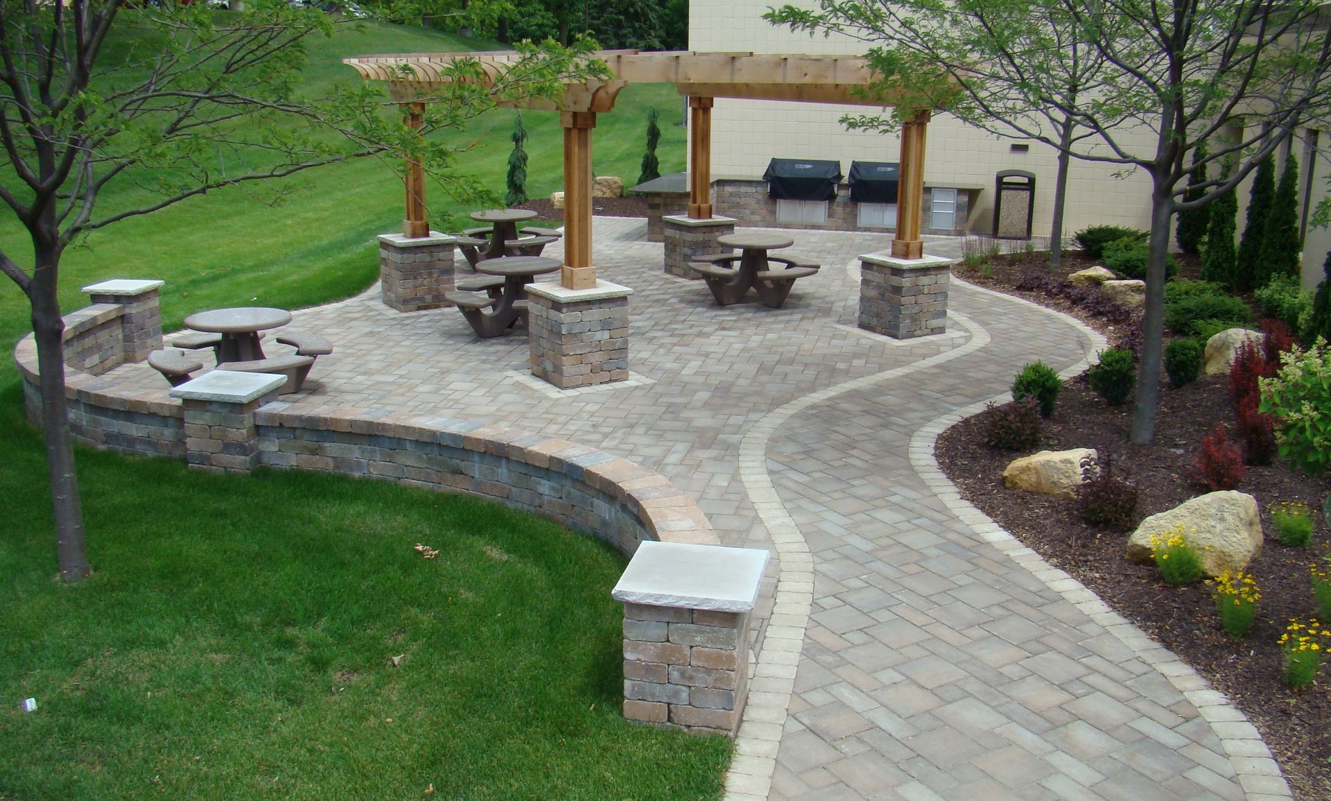 Brick patio with picnic tables, pergola, and path on a grassy hillside.