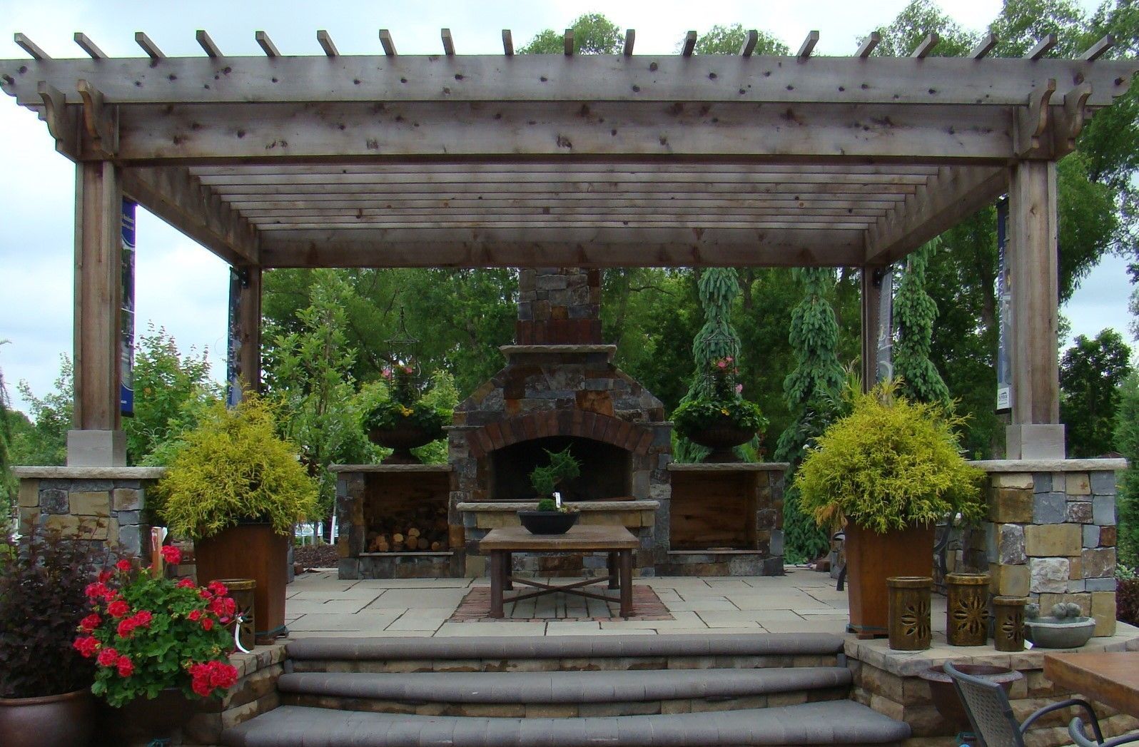 Wooden pergola with stone fireplace, patio, and potted plants in an outdoor setting.
