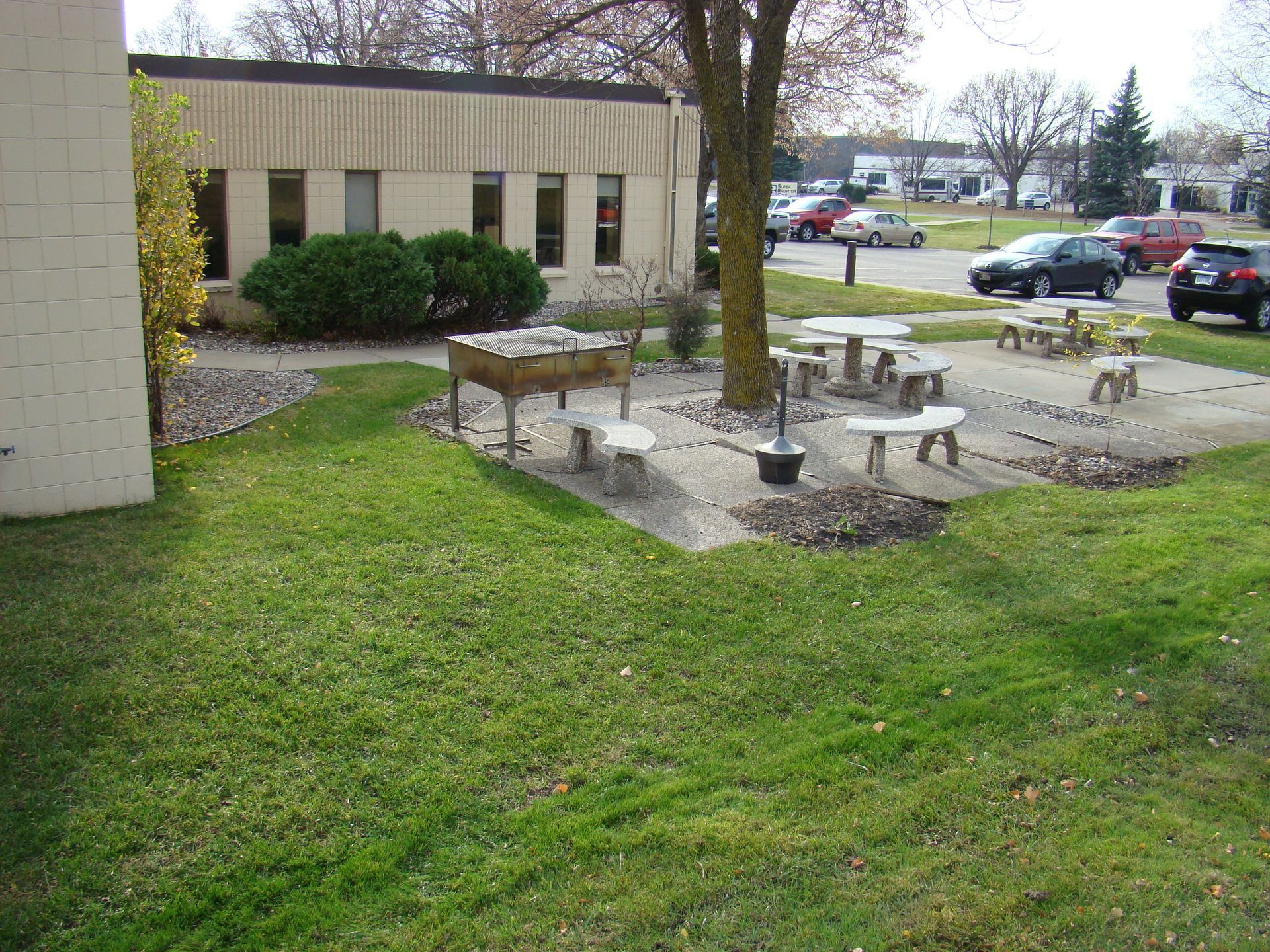 Outdoor seating area with picnic tables and a grill near a building and parking lot.