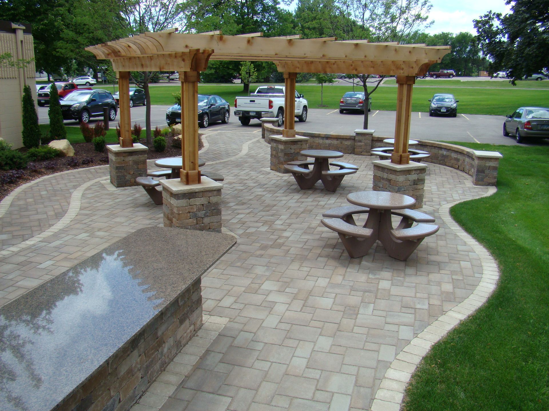 Outdoor patio with stone pavers, picnic tables, and a wooden pergola near a parking lot.