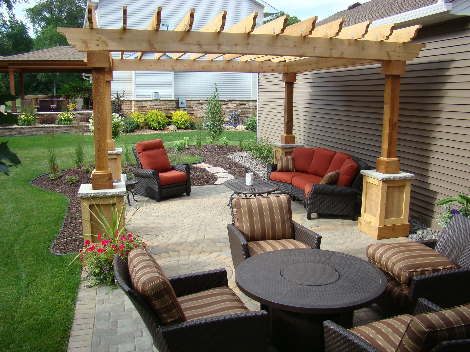 Outdoor patio with pergola, seating, and landscaping. Brown furniture, brick pavers, and lush greenery.