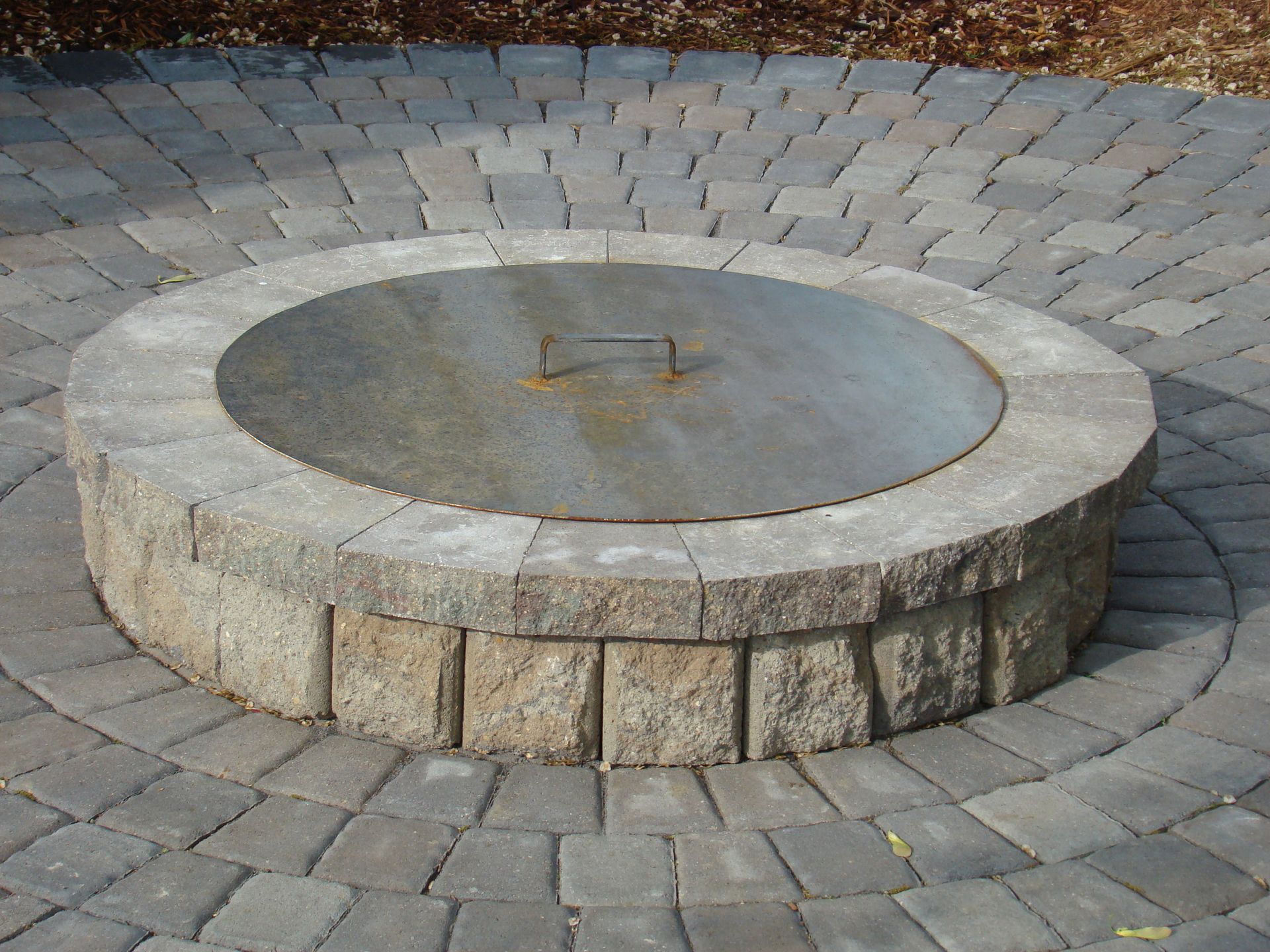 Circular stone fire pit with metal cover on a brick patio.