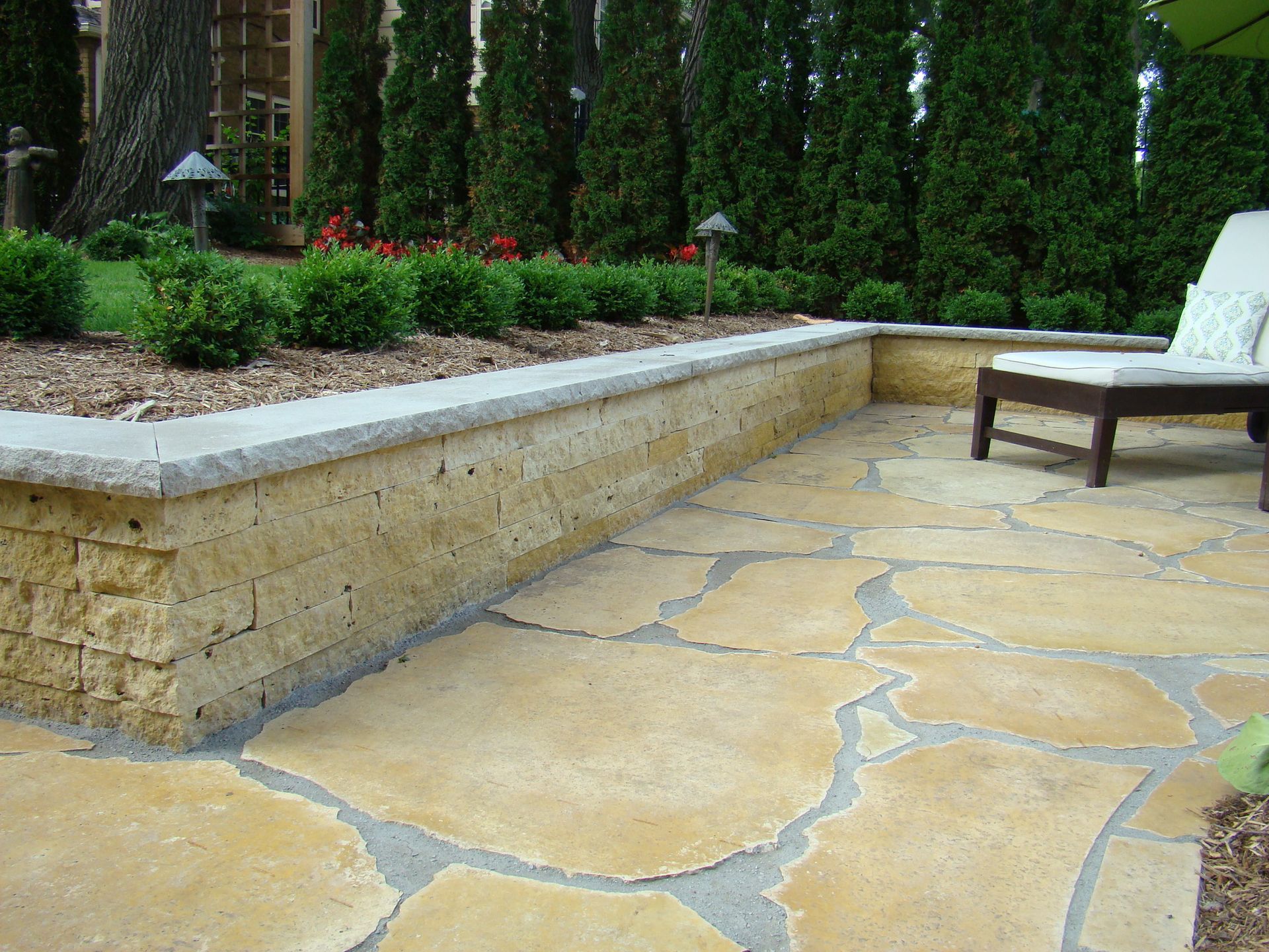 Patio with stone pavers, low stone wall, and hedge in background.