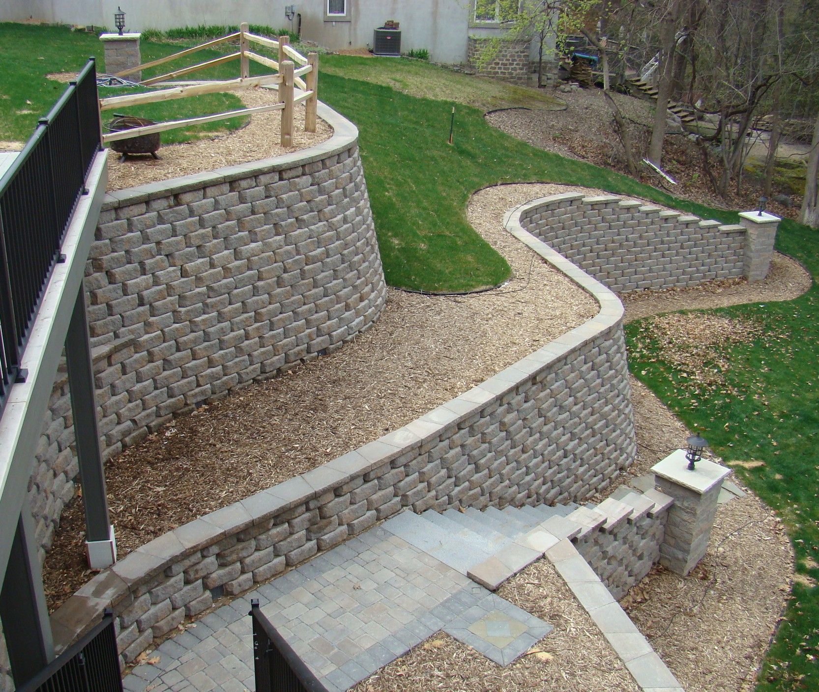 Retaining walls made of gray blocks separate levels of a landscaped yard with green grass and stone paths.
