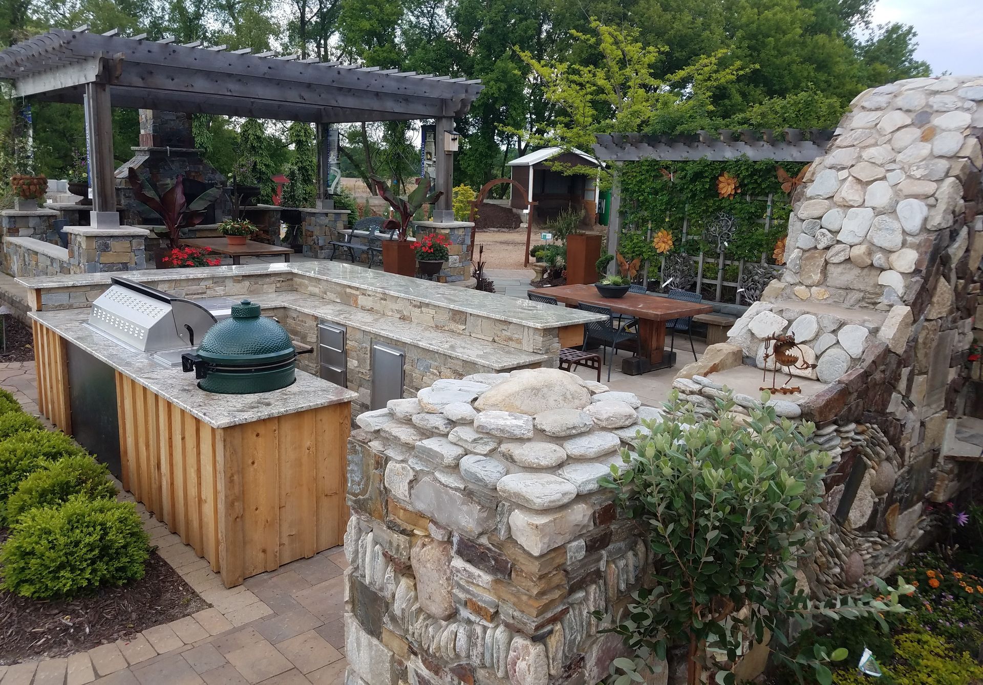 Outdoor kitchen with grill, counter space, stone and wood structure. Lush landscaping surrounds the patio.