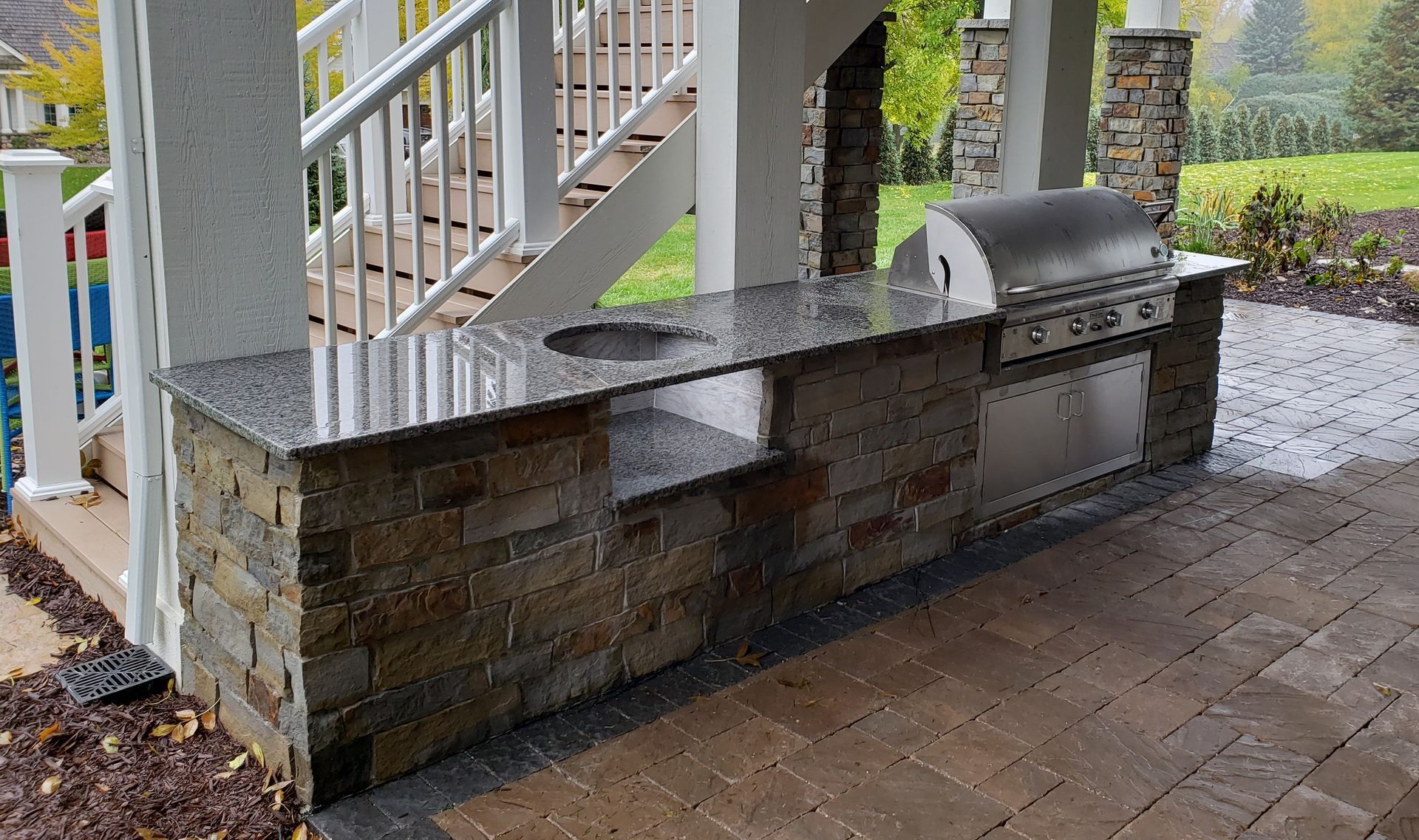 Outdoor kitchen with granite countertop, built-in grill, and stone facade, beside a deck with white railings.