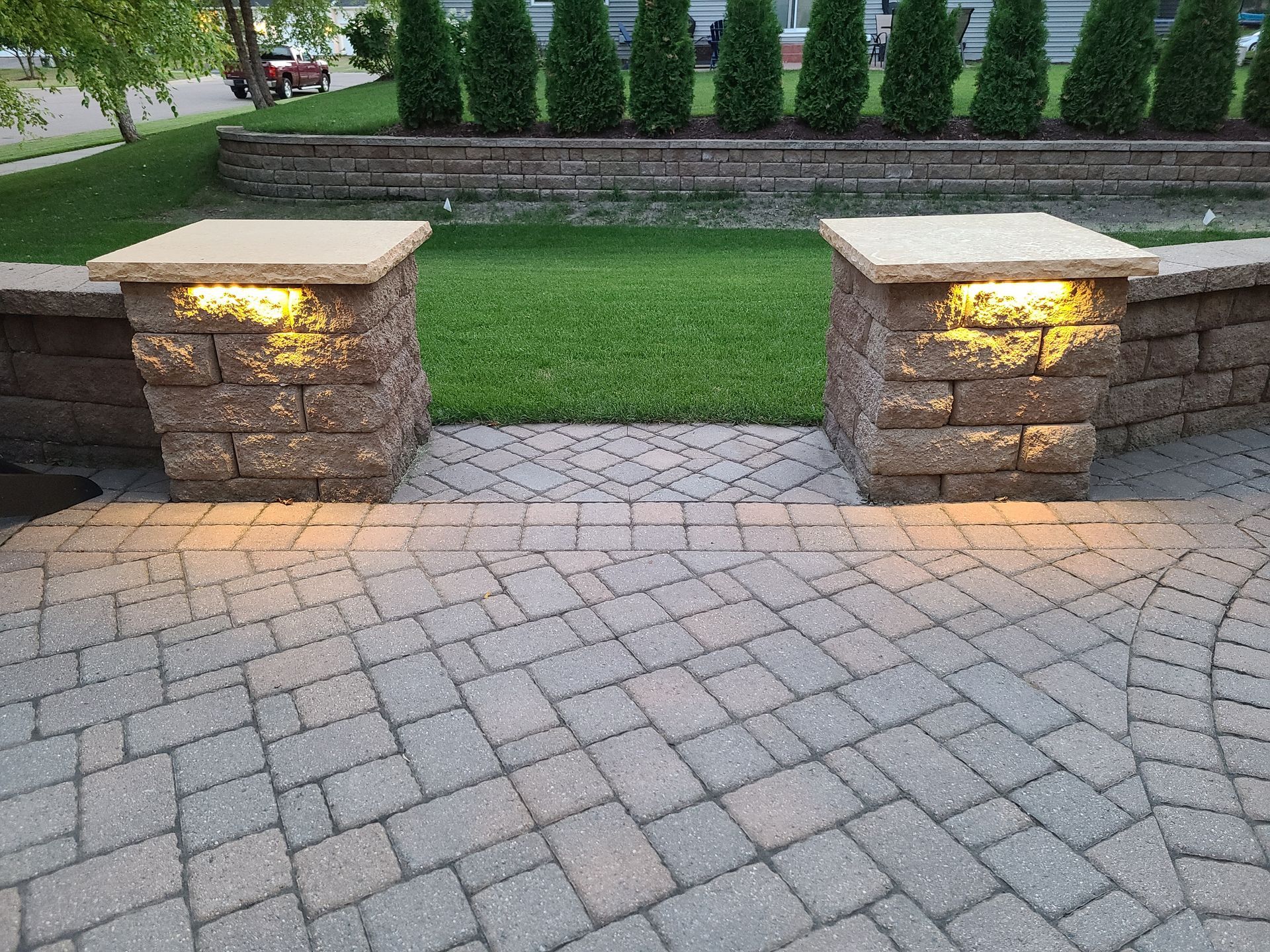 Brick pillars with lights on a paved patio, leading to a lawn with evergreen shrubs.