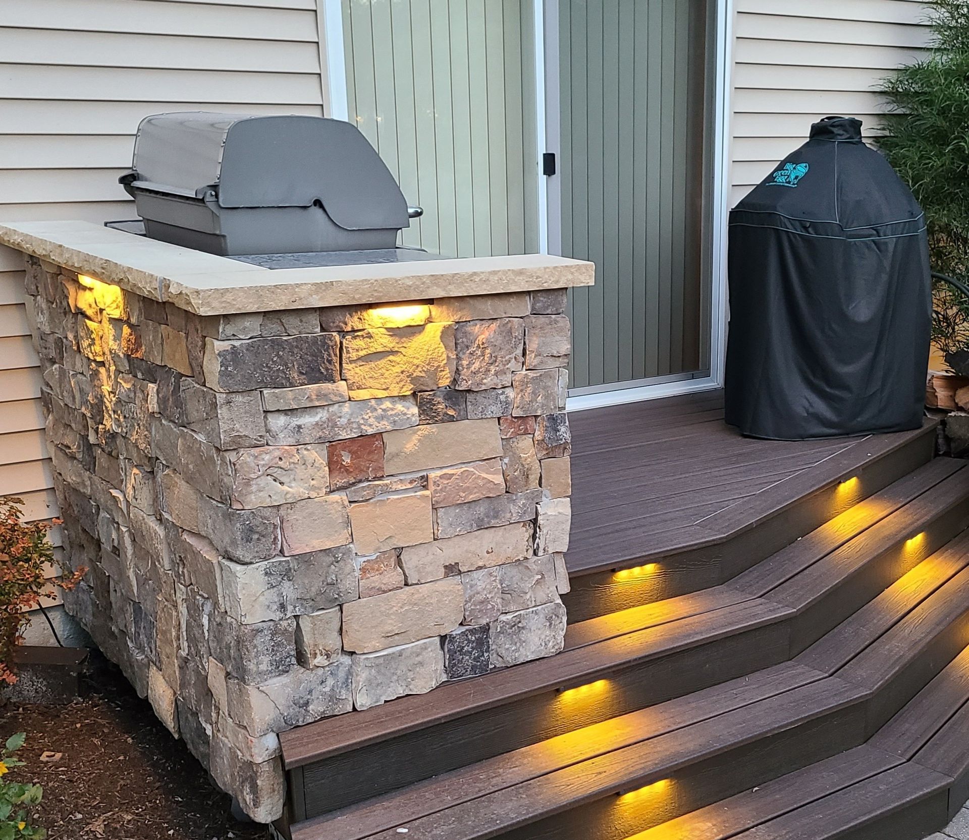 Stone outdoor kitchen with grill, deck steps, and lighting.