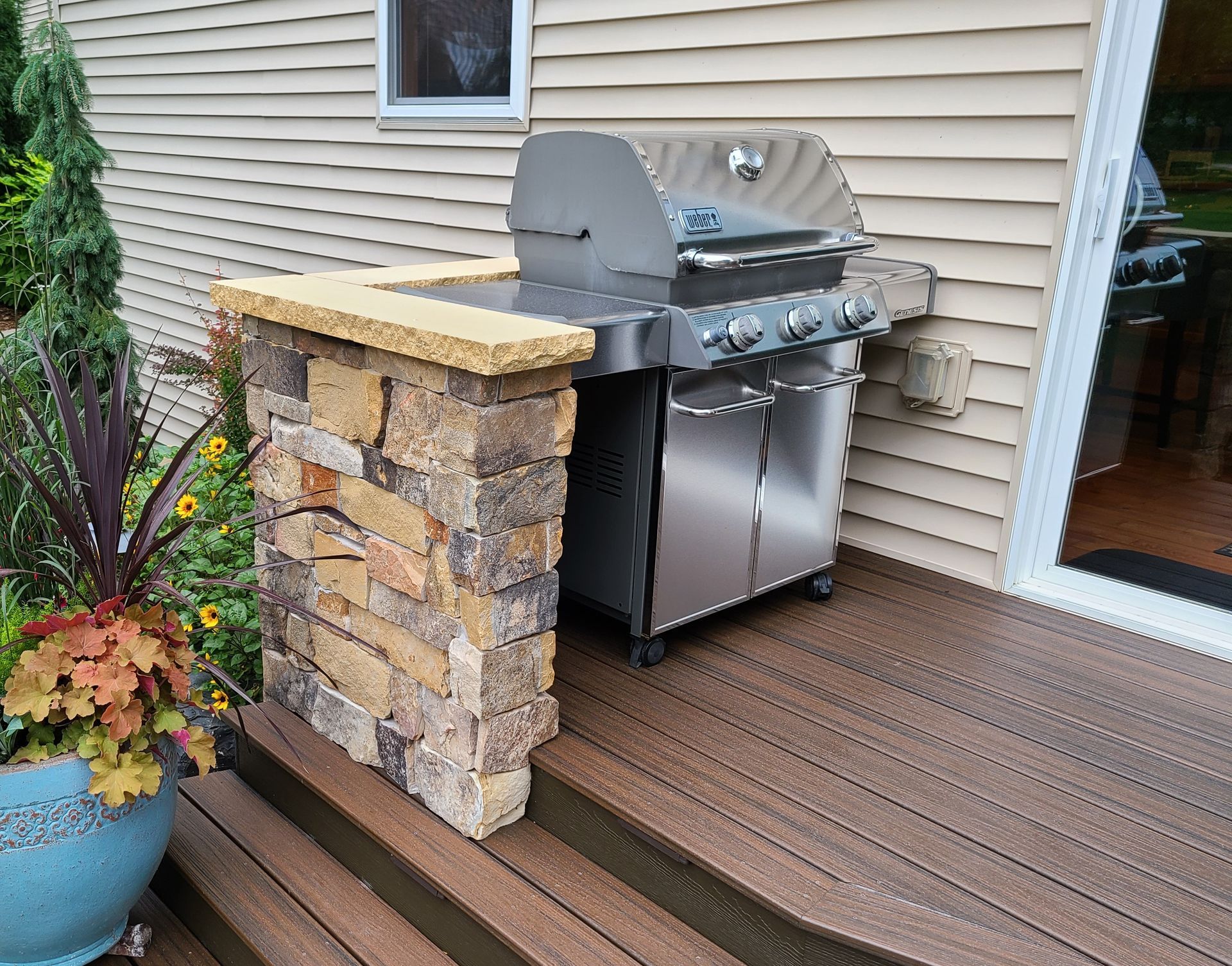 Grill station on a deck, with a brick pillar and countertop, next to the house with a sliding door and potted plants.