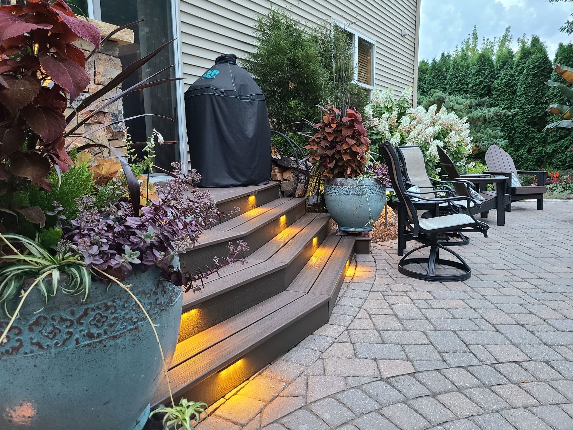 A patio with steps, outdoor furniture, and potted plants lit with warm lighting. A grill is covered near steps.