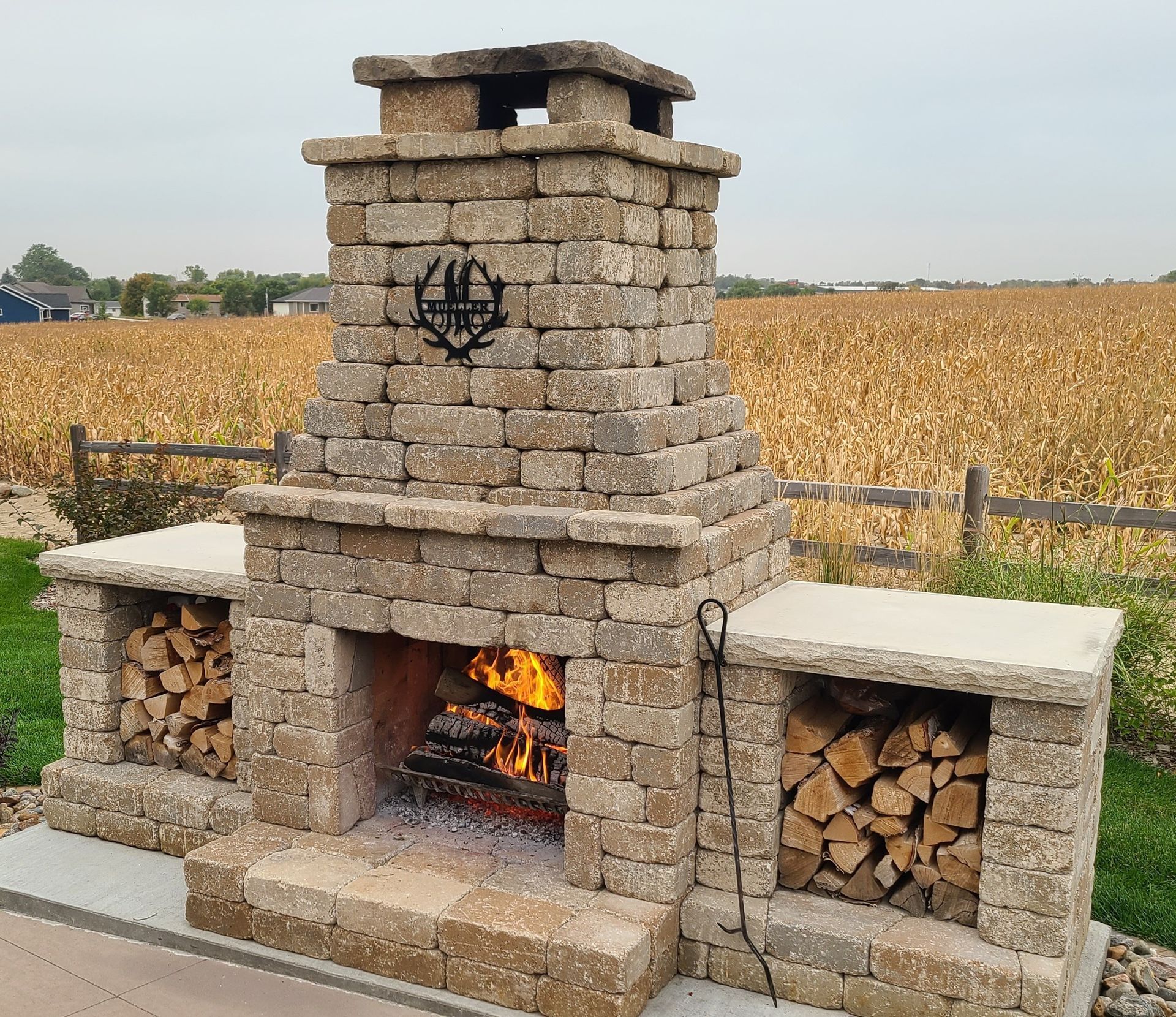 Stone outdoor fireplace with burning fire and wood storage, set against a cornfield.