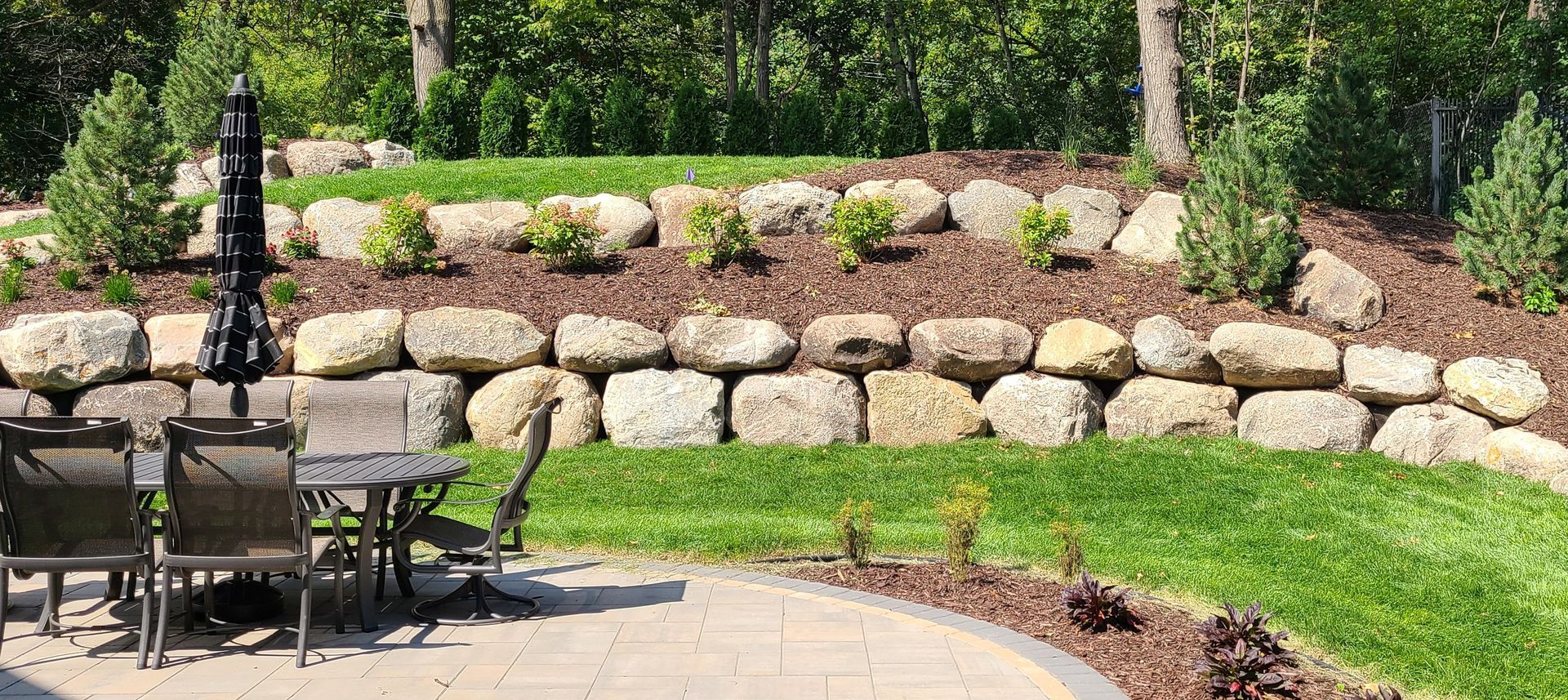 A tiered garden with stone retaining walls, plants, grass, and a patio with outdoor furniture.