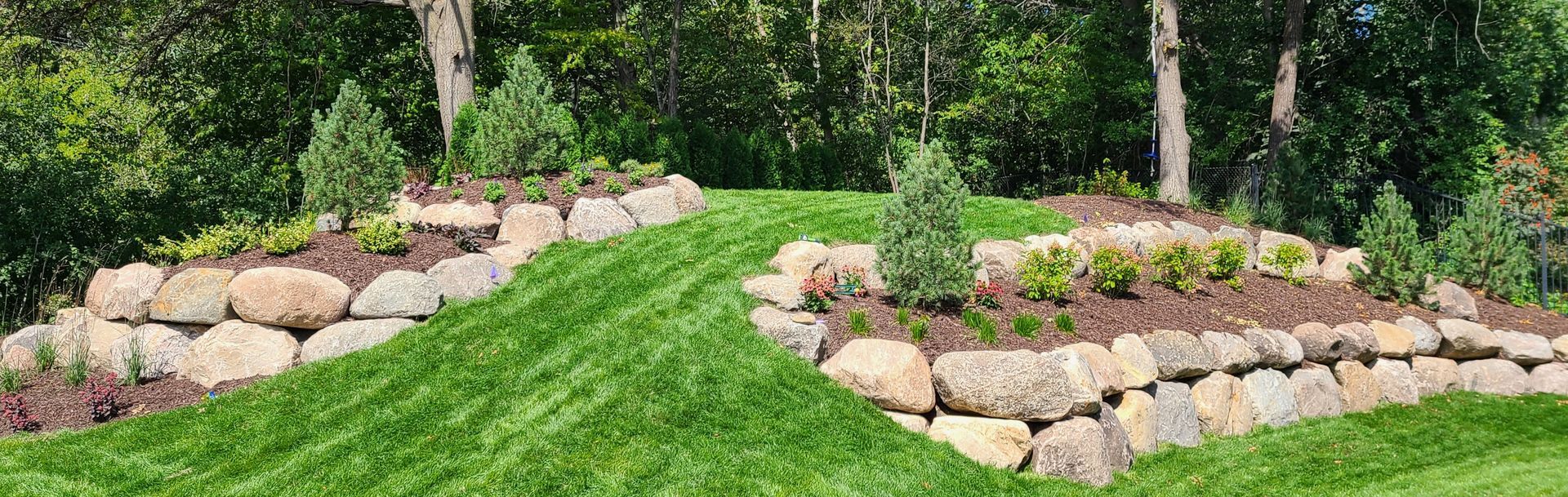 Green grass hillside with stone retaining walls and plantings. Trees are in the background.