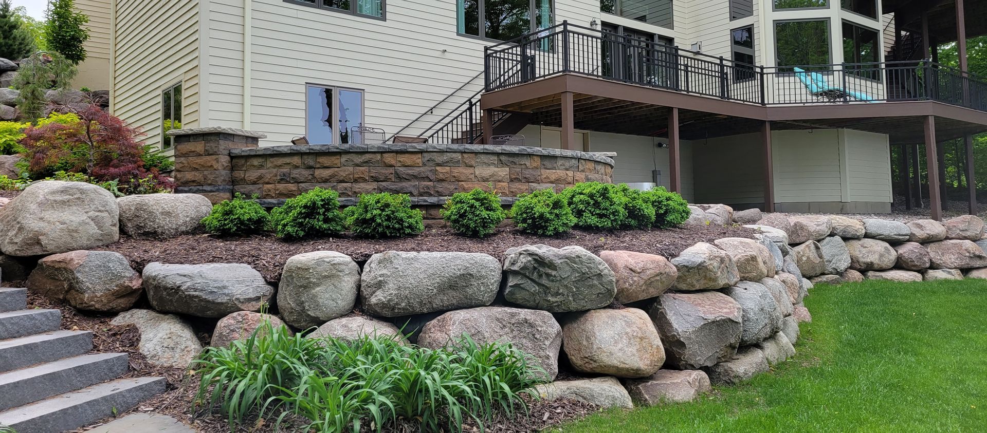 Large stone retaining walls in front of a house, with green shrubs, lawn, and a deck.