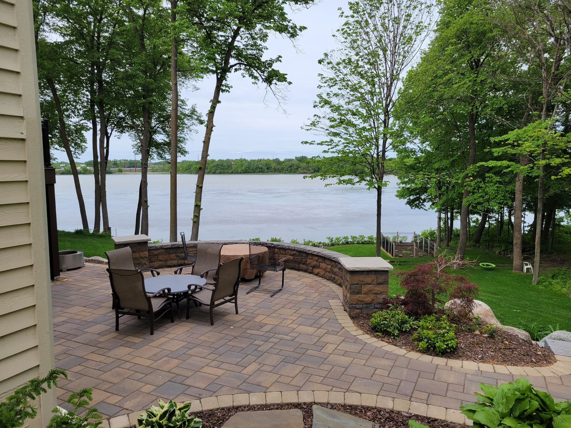 Patio overlooking lake, with seating and landscaping. Brick pavers, trees, and cloudy sky.