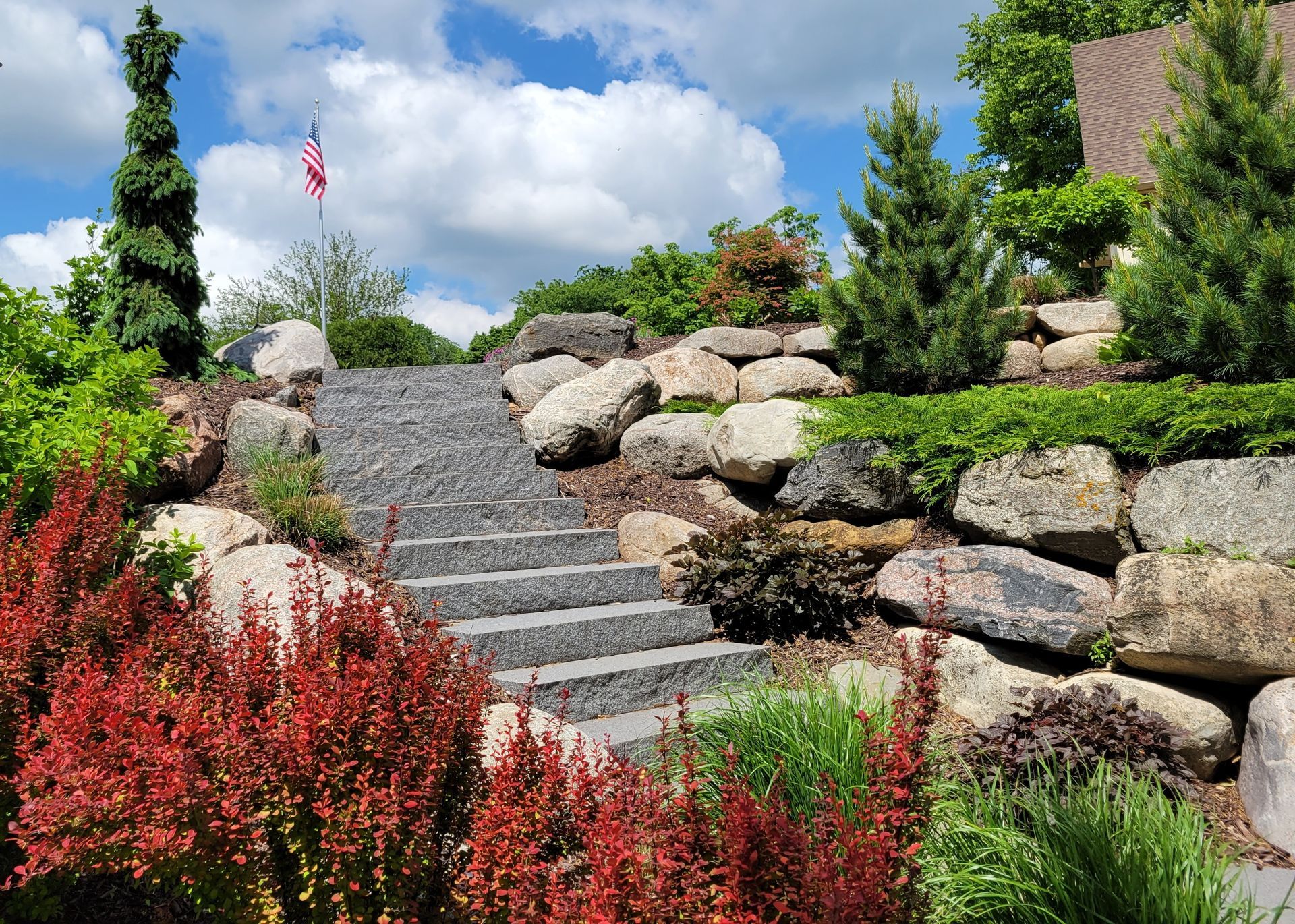 Stone steps ascend a landscaped hillside with colorful shrubs, rocks, and trees under a blue sky.