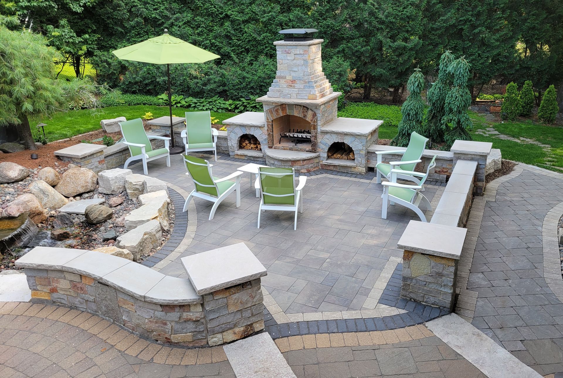 Outdoor patio with fireplace, seating, and waterfall feature. Green chairs around fireplace, umbrella. Stone and brick construction.
