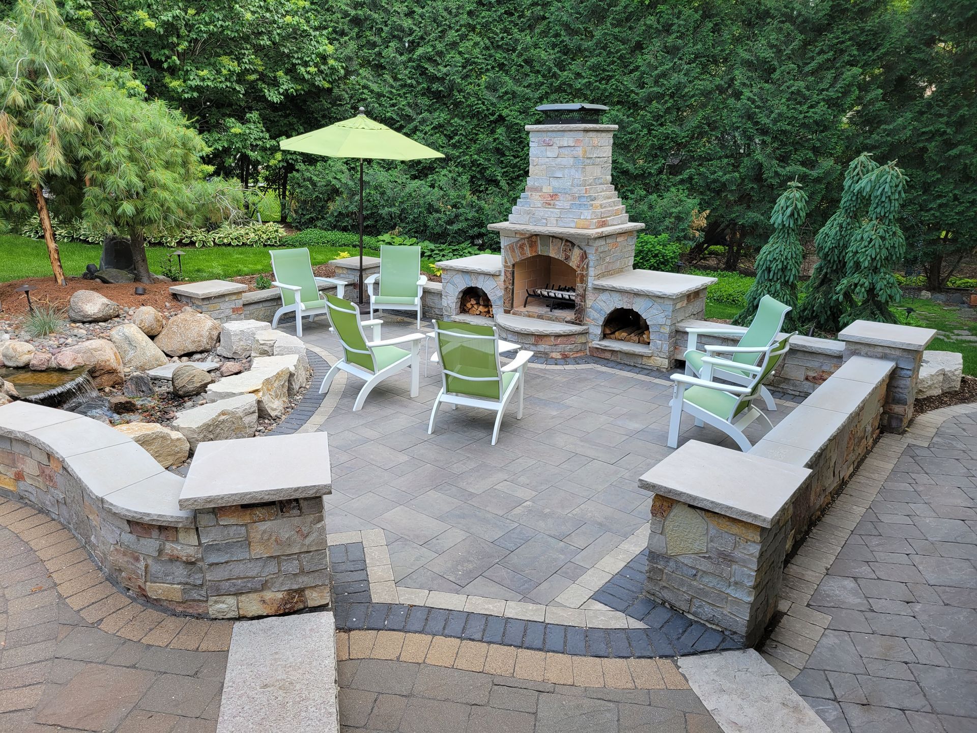 Stone patio with fireplace, green chairs around a fire pit, and small pond.