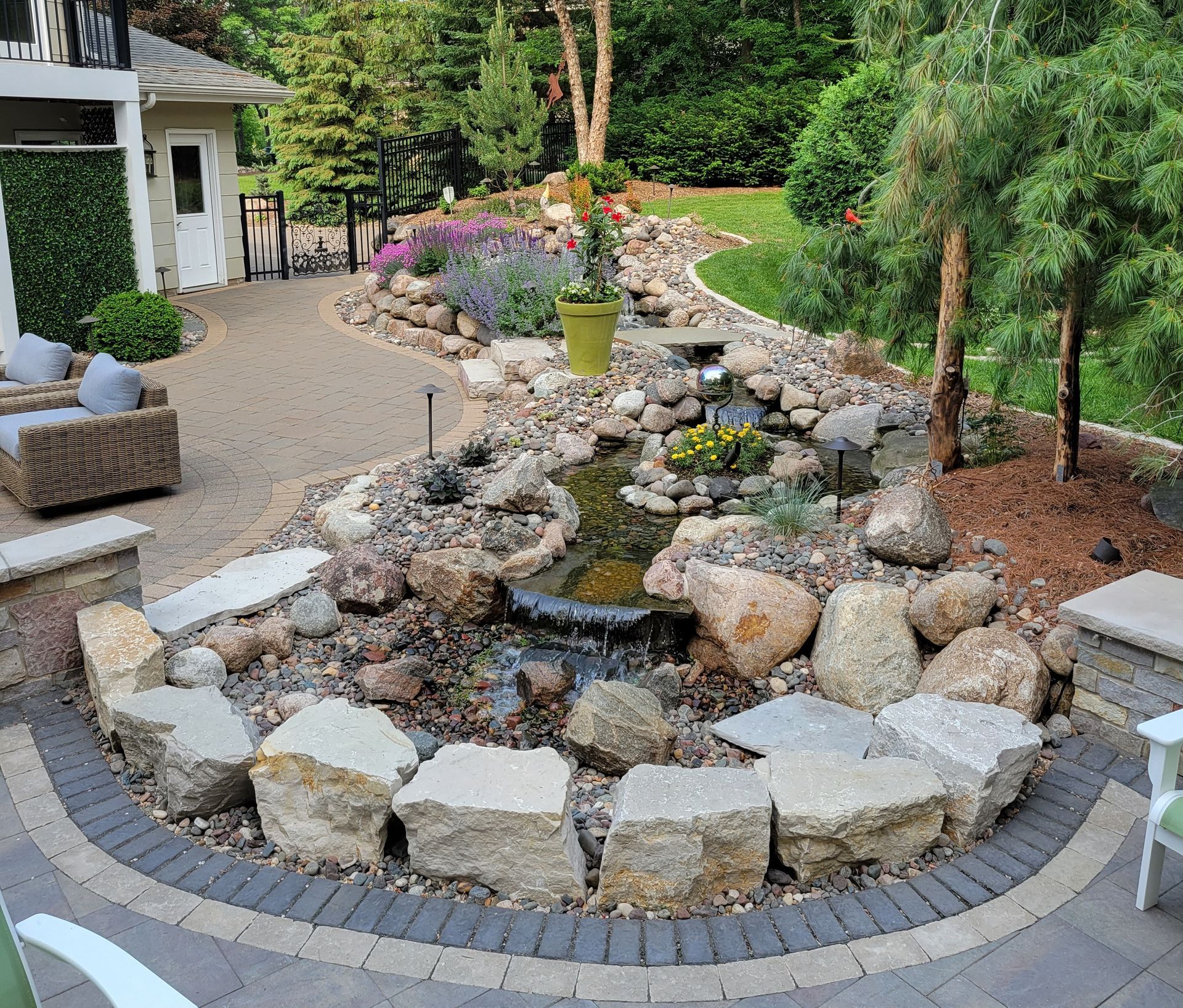 A backyard pond with waterfall surrounded by large rocks, plants, and a paver patio.