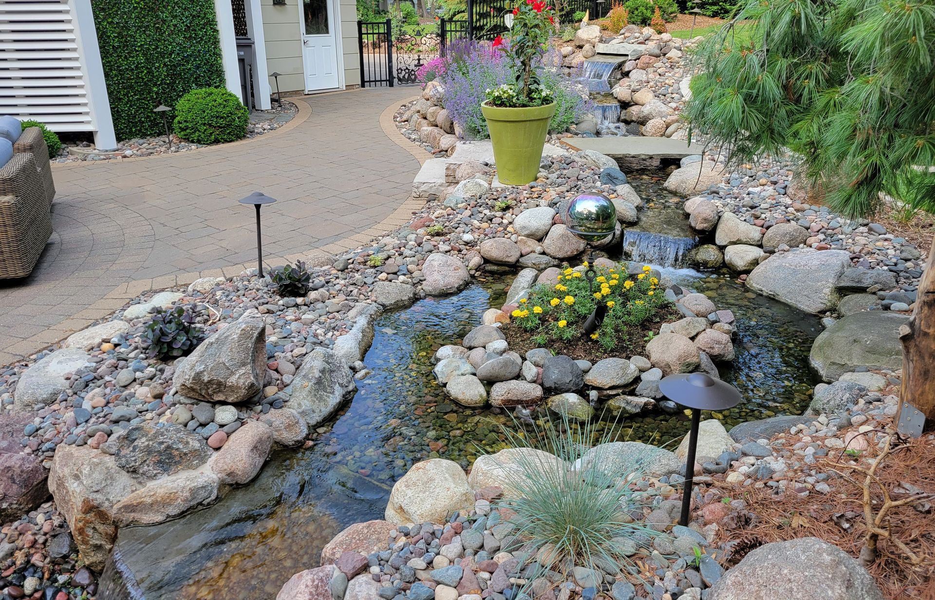 A winding water feature with a stone border, plants, and a spherical fountain in a garden.