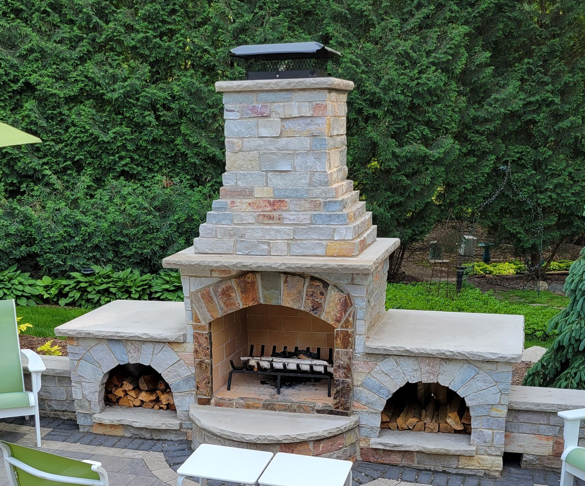 Outdoor stone fireplace with built-in wood storage. Archway firebox, square chimney, set on patio with greenery backdrop.