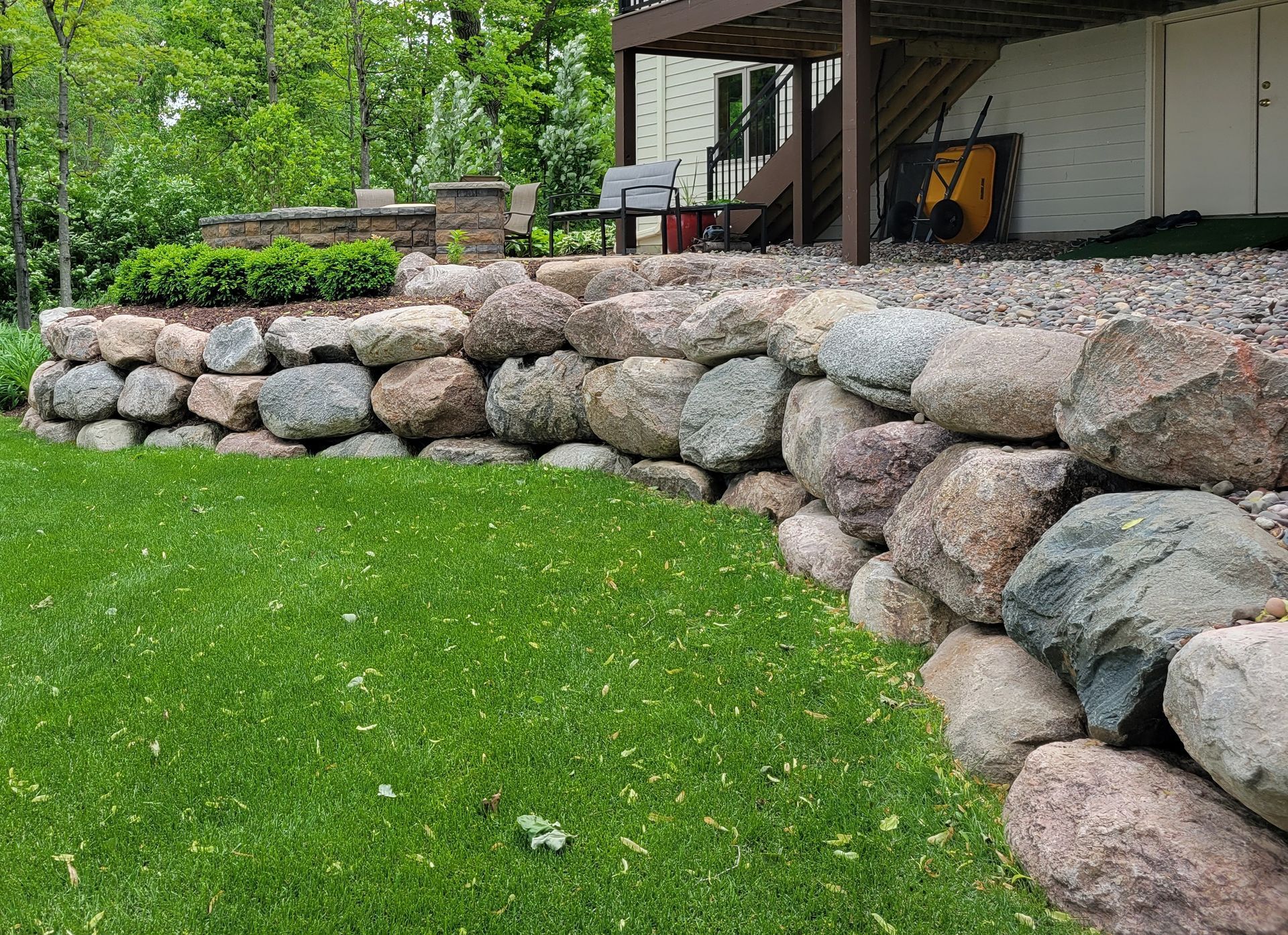 Stone retaining wall bordering a green lawn, leading to a deck and house.