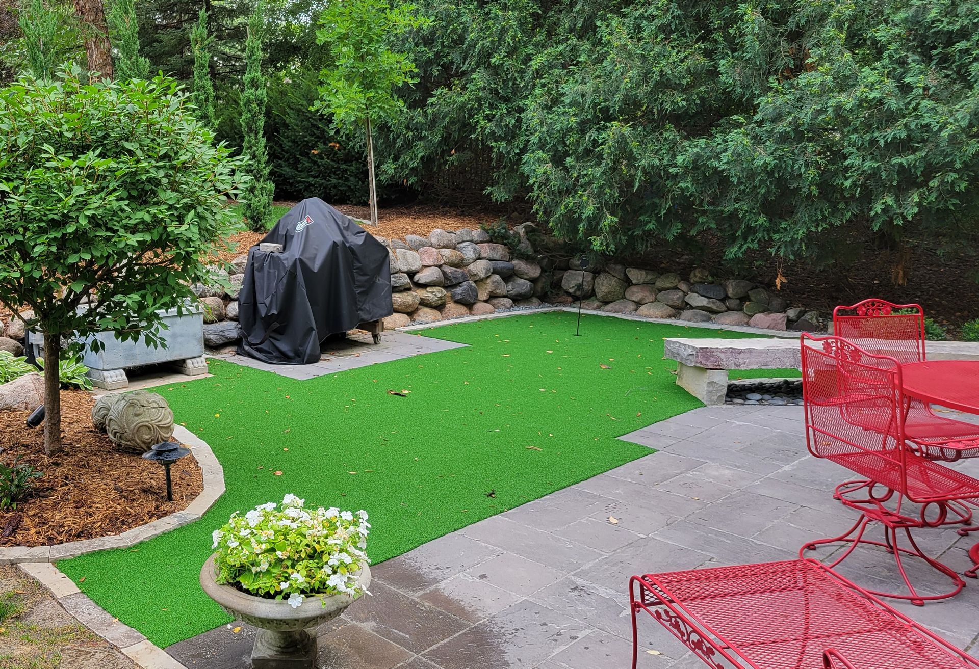 Backyard patio with green turf, stone wall, grill, and red patio furniture.