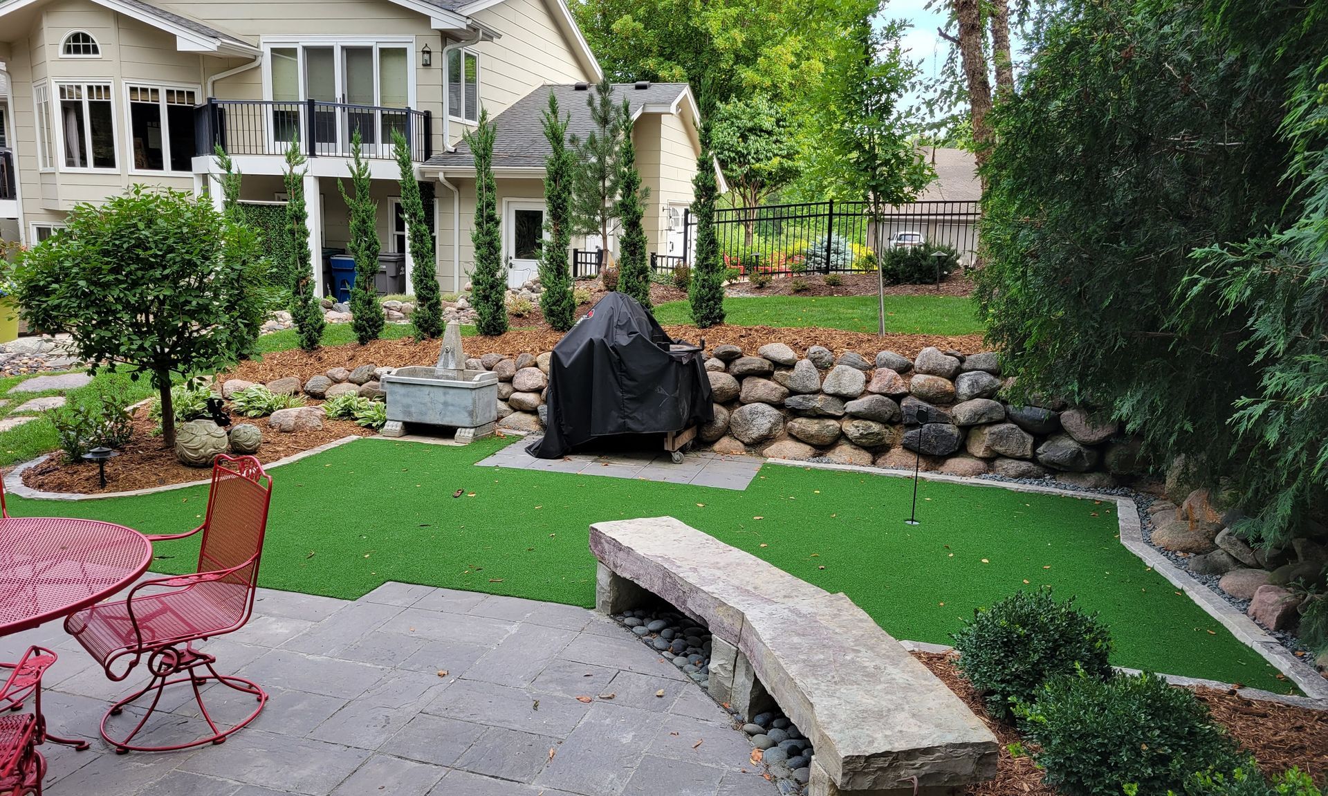 Backyard with green turf, stone retaining wall, patio furniture, and a grill.