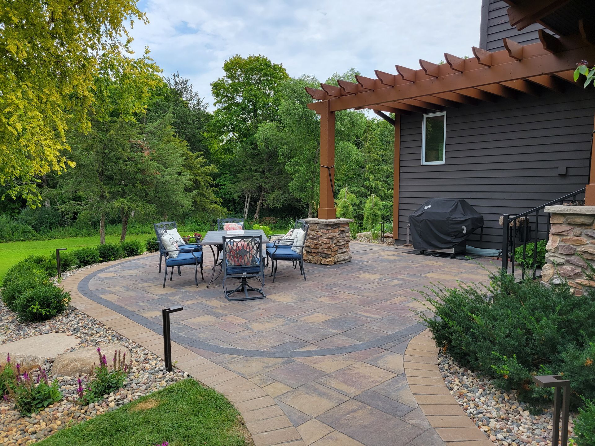 Patio with stone pavers, seating, fire pit, and pergola next to a house with dark siding.
