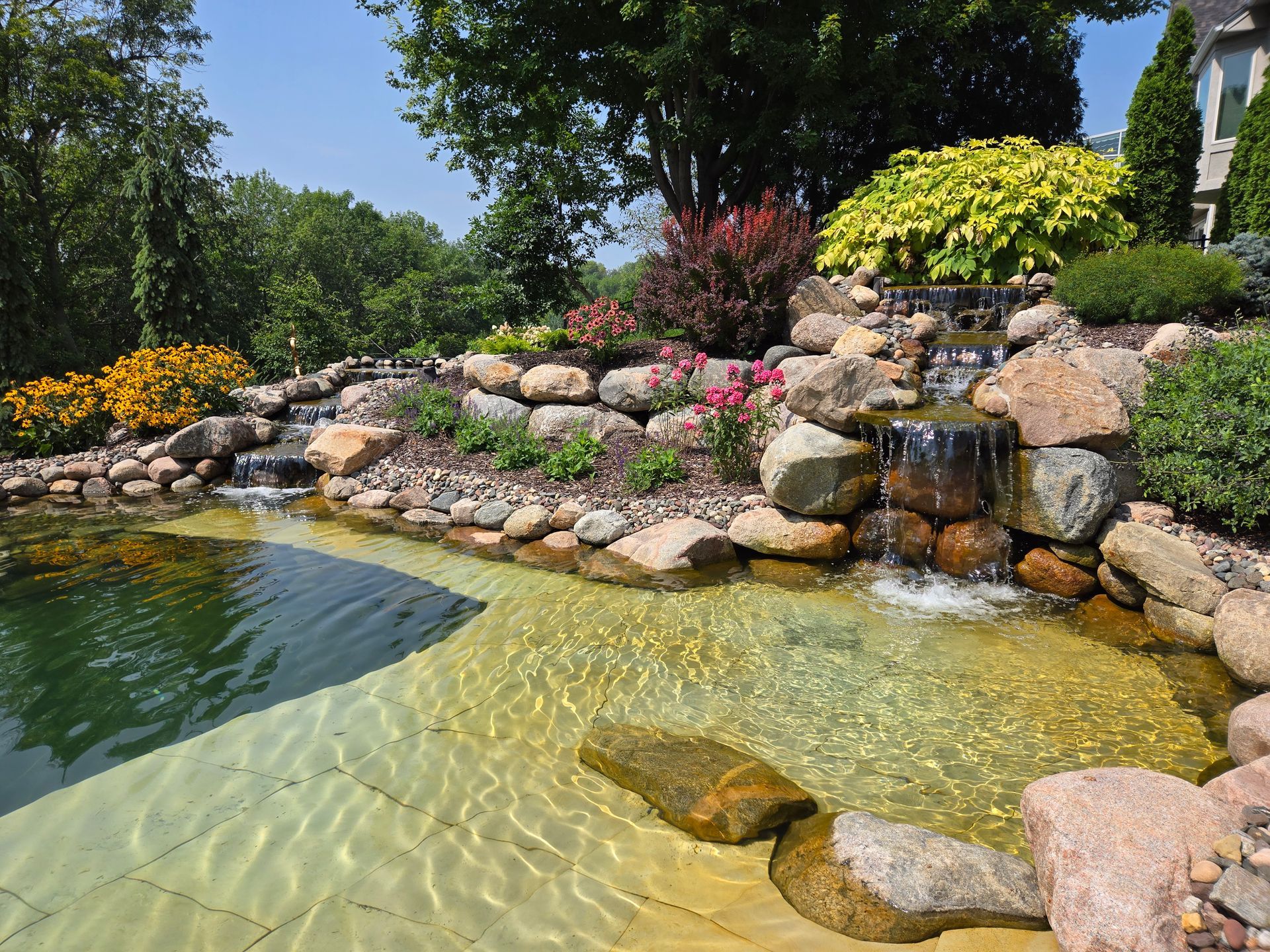 Stone waterfall cascading into a pond with landscaped rocks and colorful plants under a blue sky.