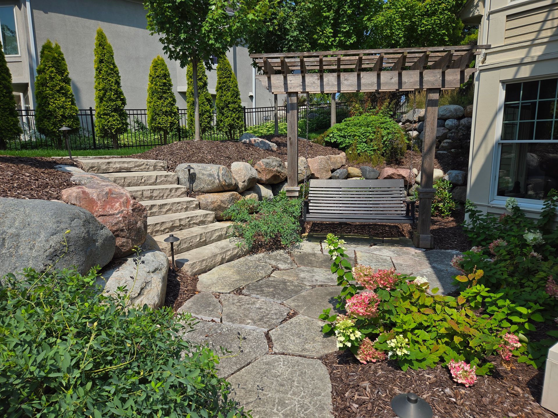 Stone steps and pathway lead to a bench under a pergola in a landscaped garden with rocks and greenery.