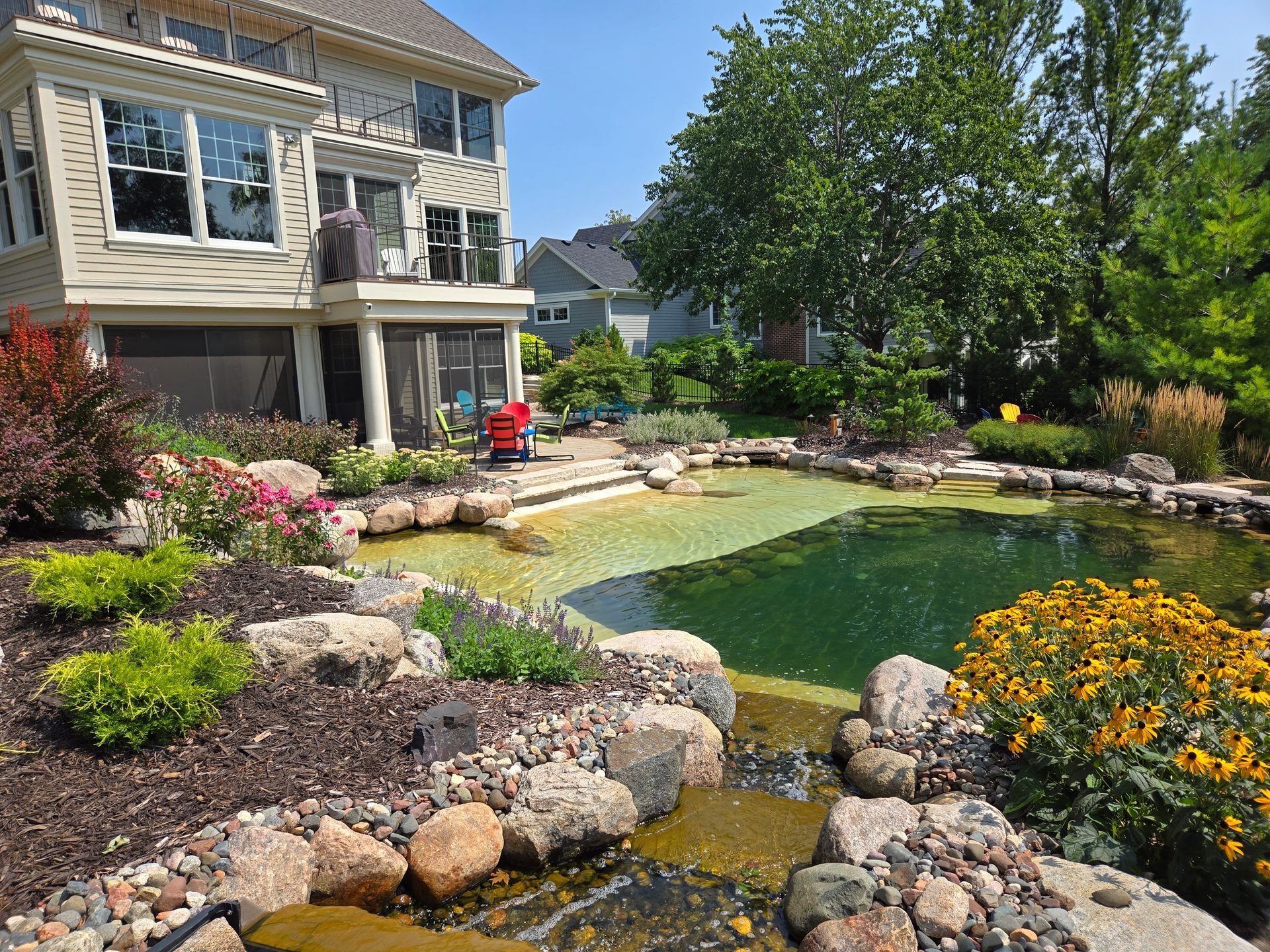 Backyard with a pond, lush landscaping, and a two-story beige house. Sunny day with blue sky.