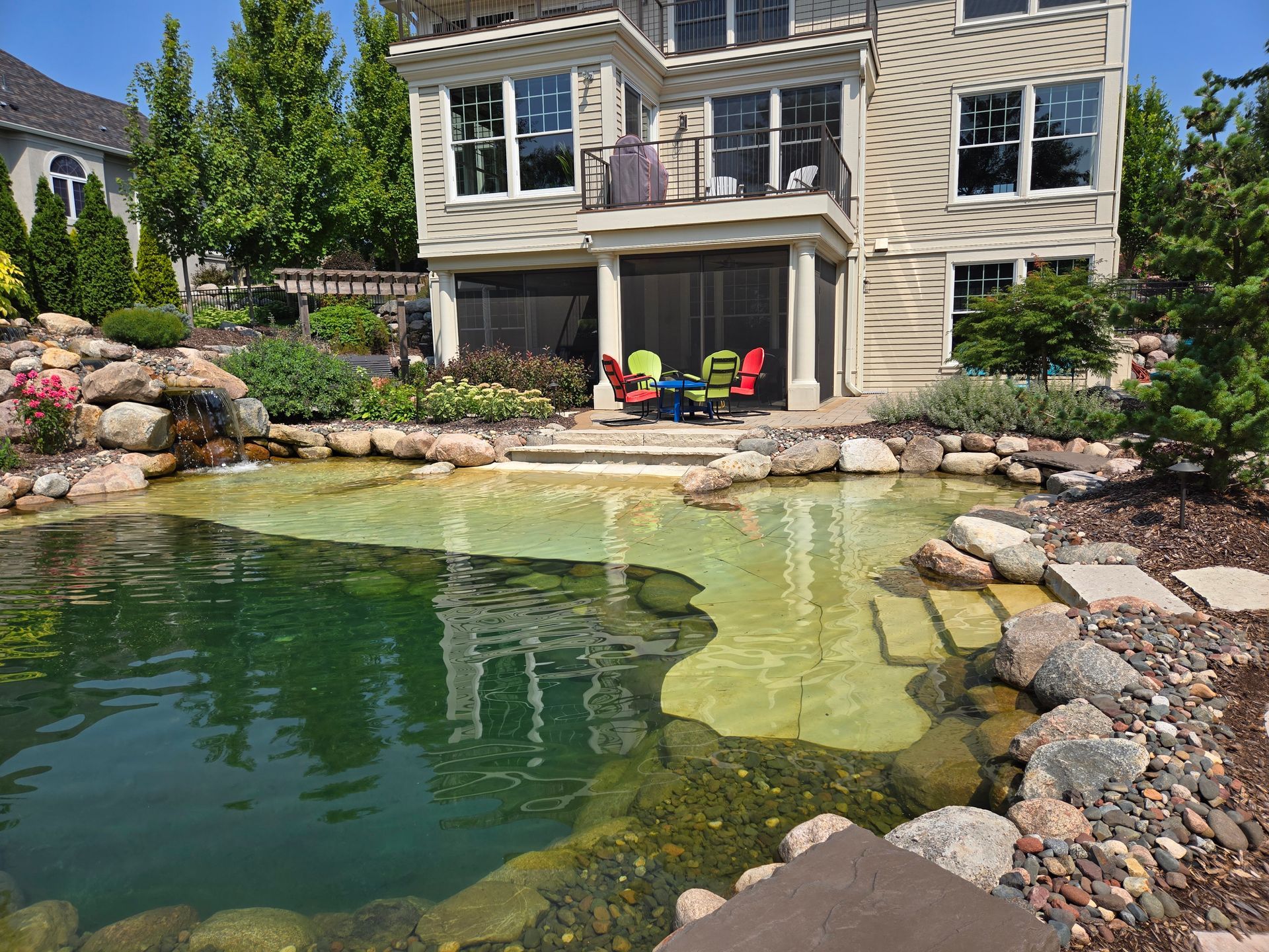 Backyard with a house overlooking a pond. The pond has rocks, stairs, and a shallow area.
