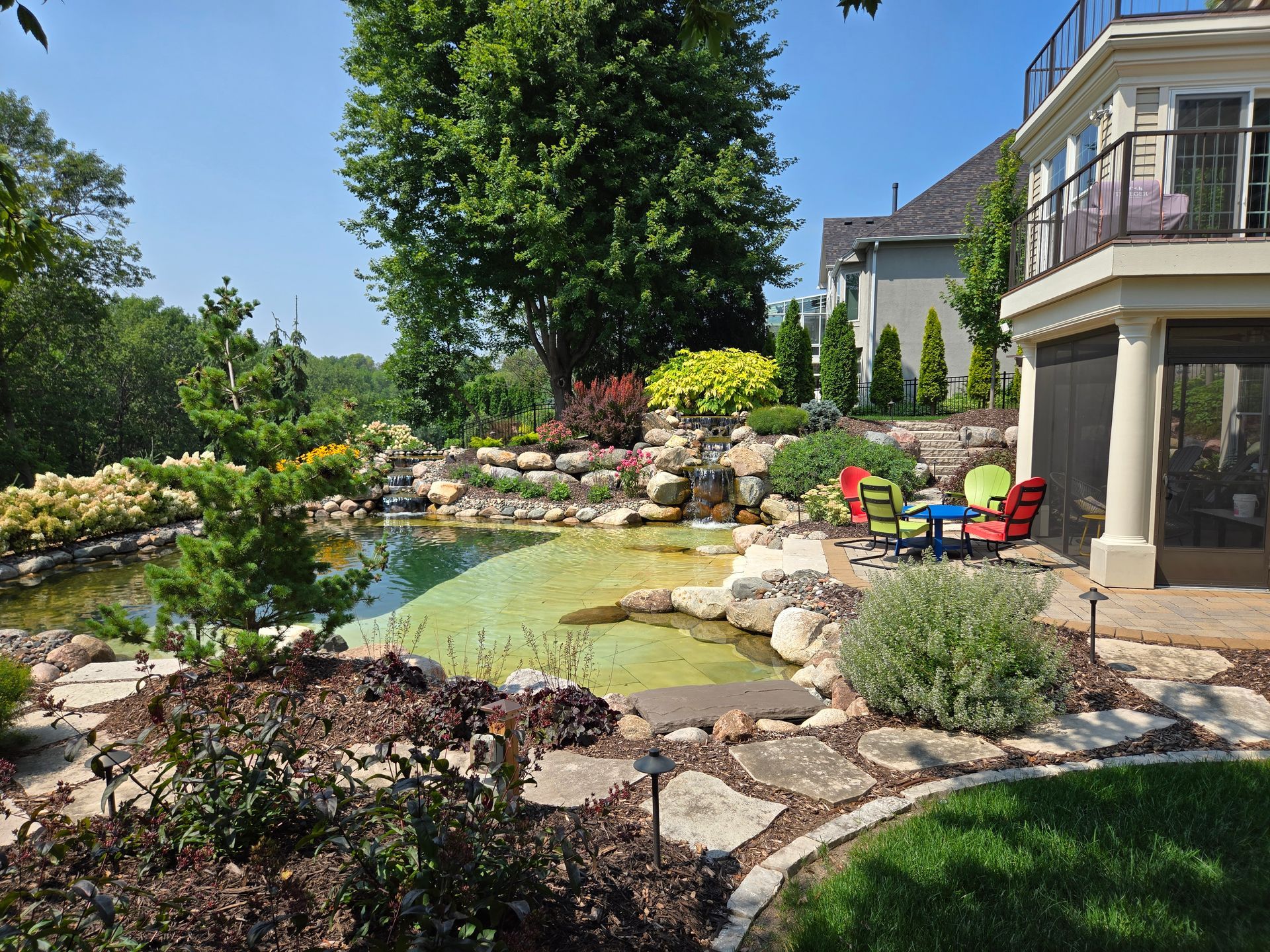 Backyard landscape with pond, stone pathway, colorful flowers, and house.
