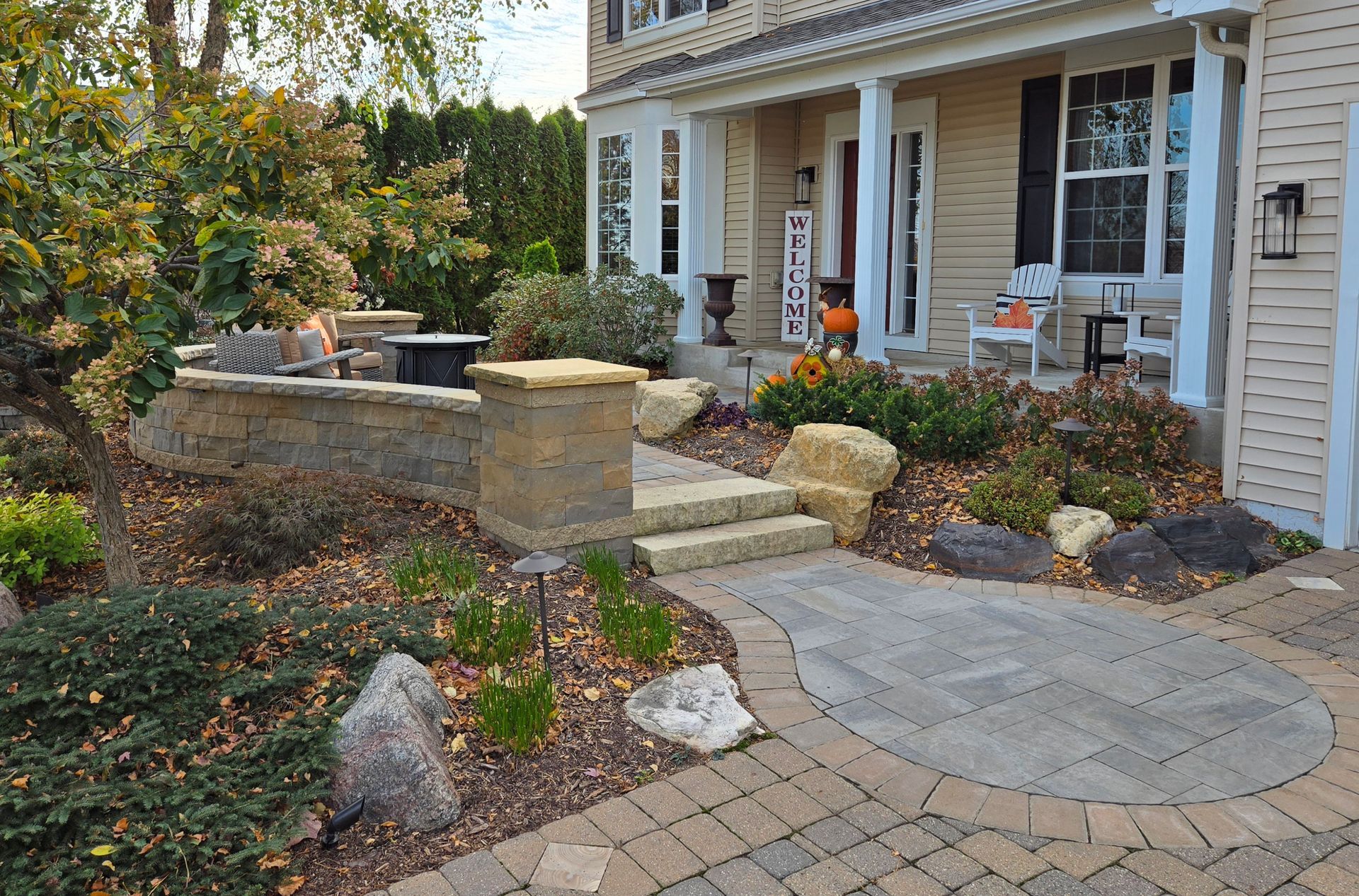 Stone walkway leads to a beige house with a porch and landscaping. Includes a fire pit and fall decor.