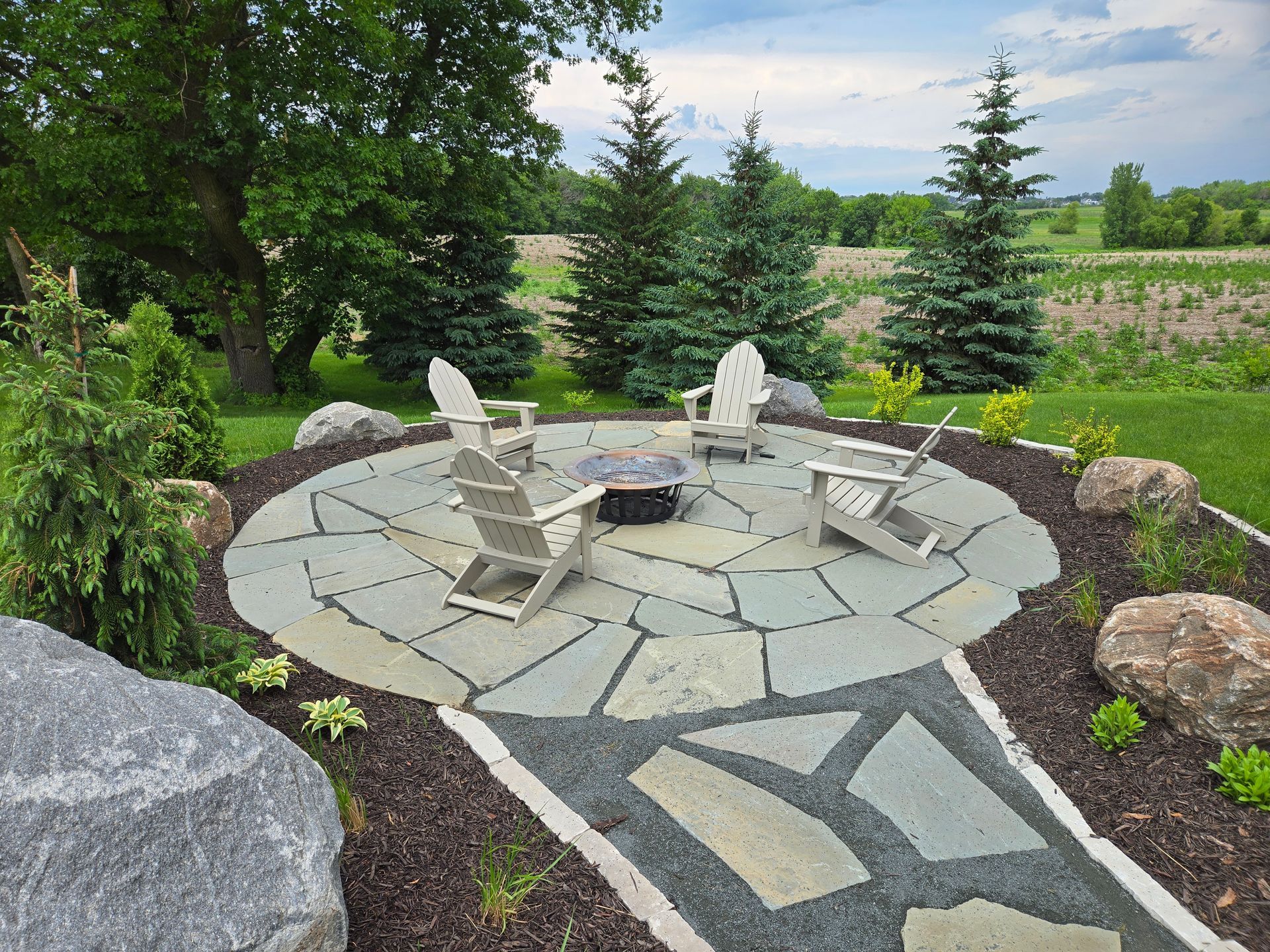 Stone patio with Adirondack chairs around a fire pit, surrounded by landscaping and trees.