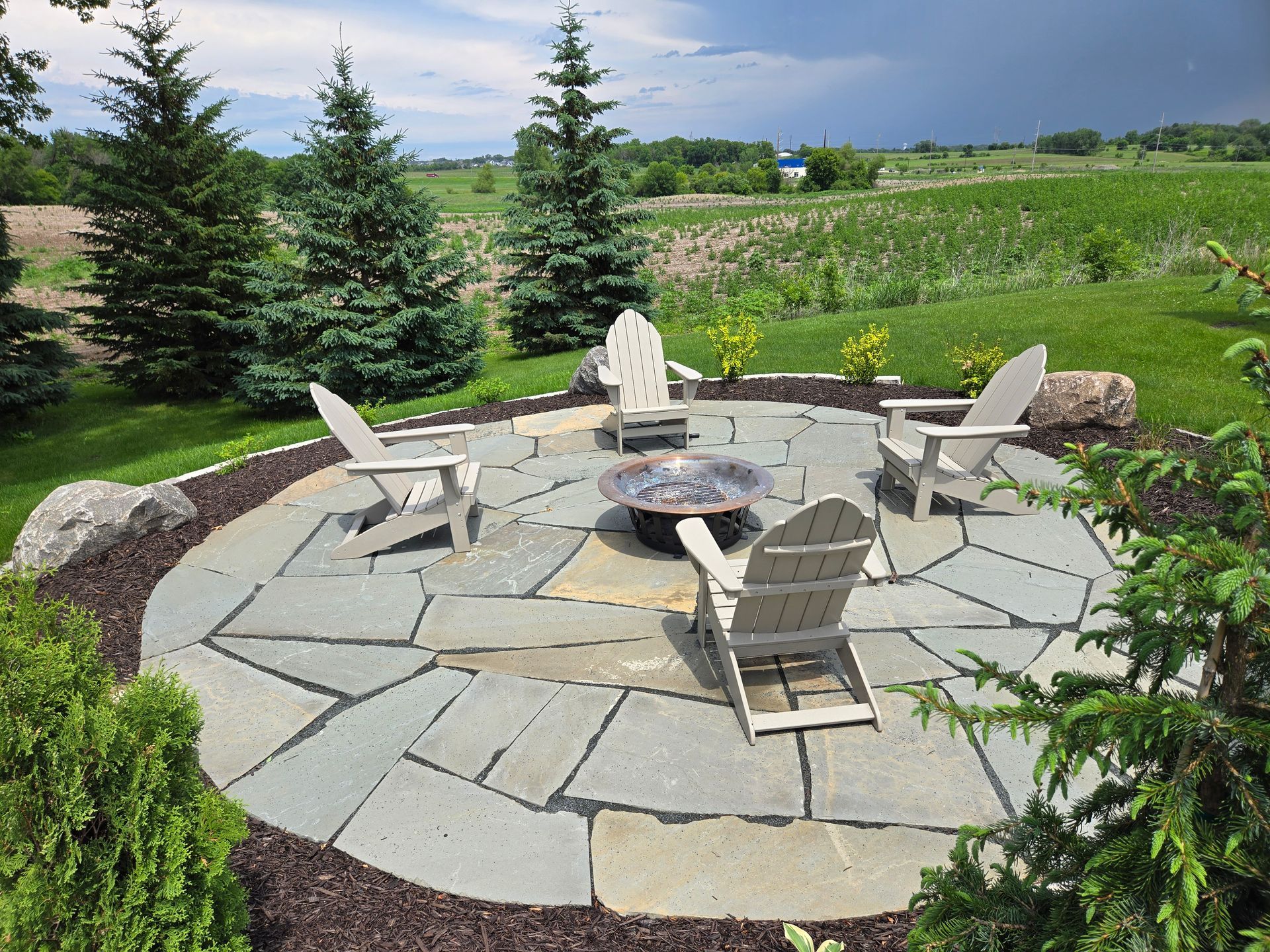 Round stone patio with Adirondack chairs around a fire pit, surrounded by plants and a field.