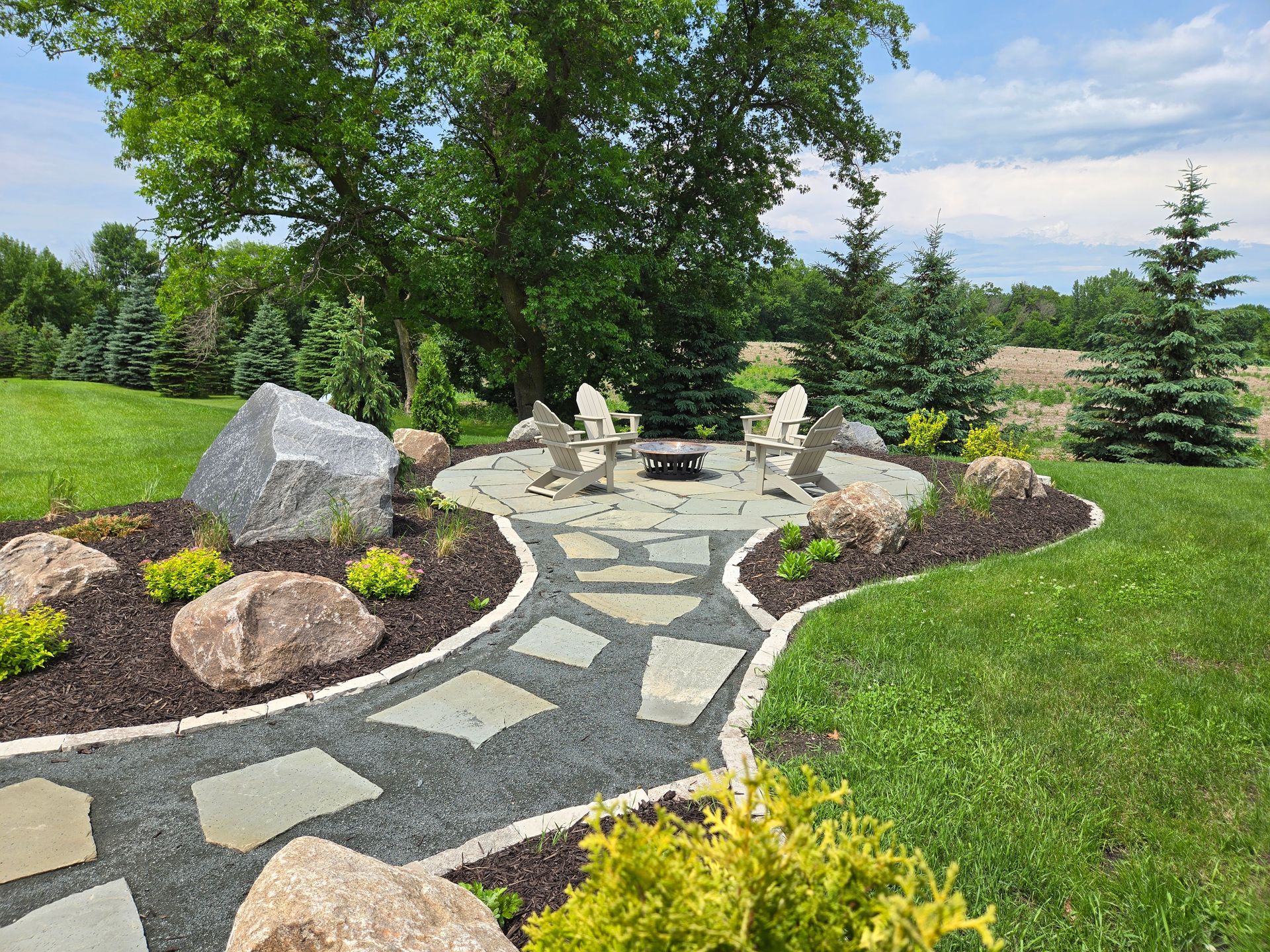 Stone patio with fire pit, stone path, chairs, boulders, and trees in a grassy yard.