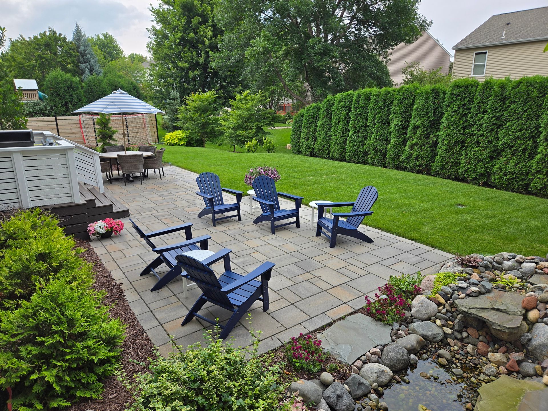 Backyard patio with blue Adirondack chairs on brick pavers, lawn, and greenery.