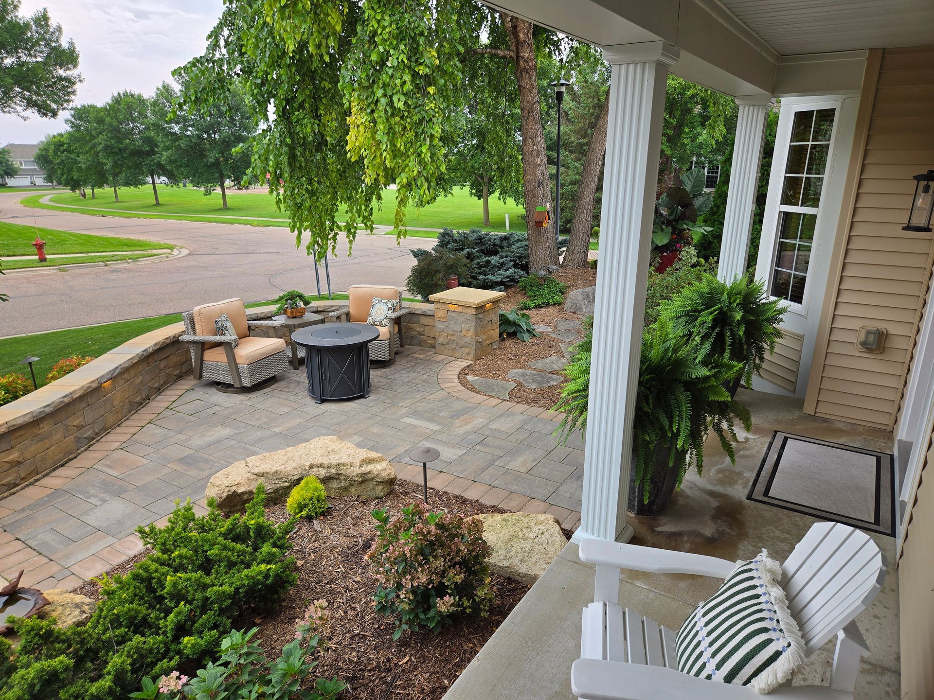Outdoor patio with seating, fire pit, and landscaping, beside a house with a front porch.
