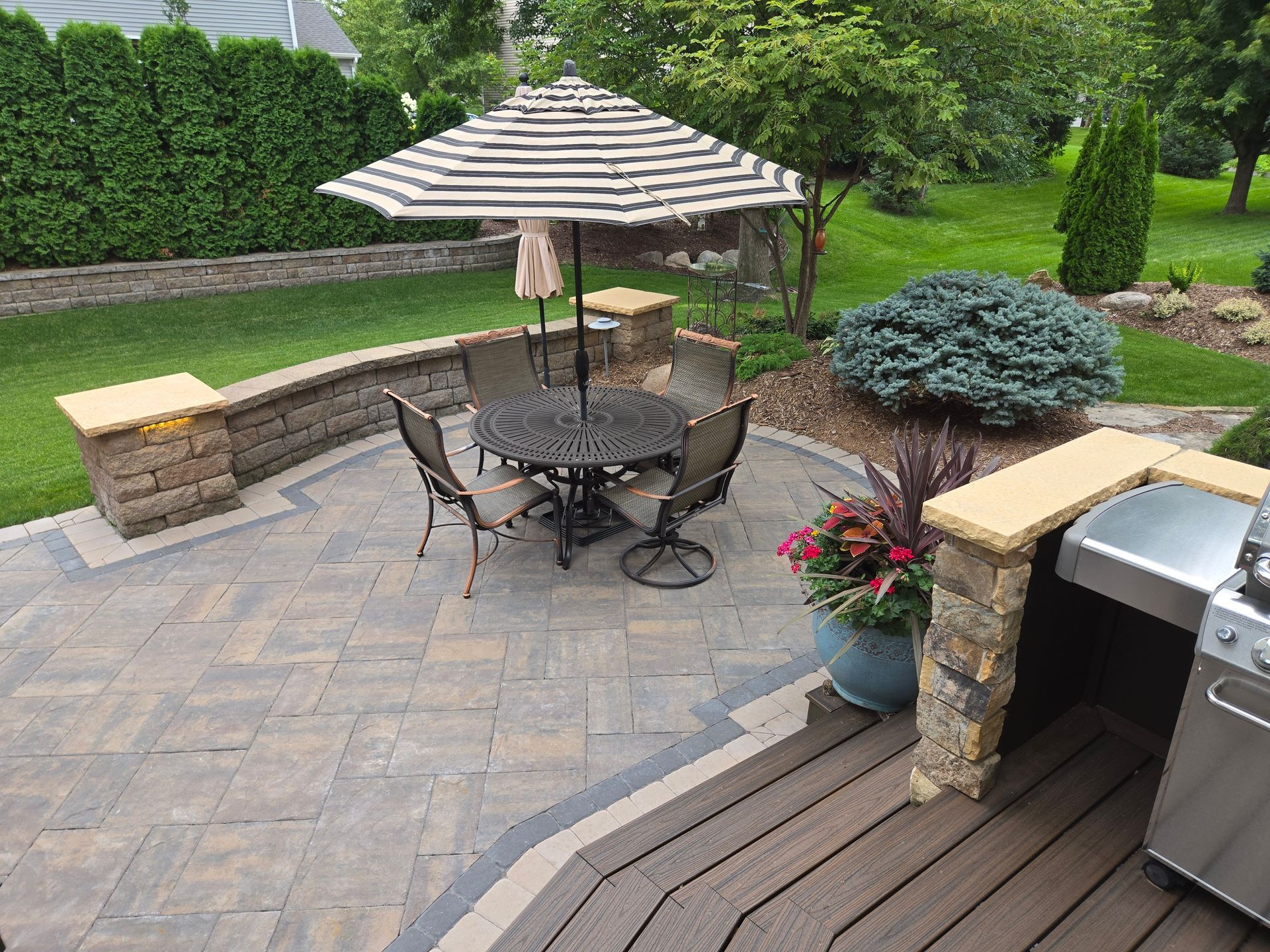 Patio with outdoor furniture, umbrella, and grill surrounded by landscaping and lawn.