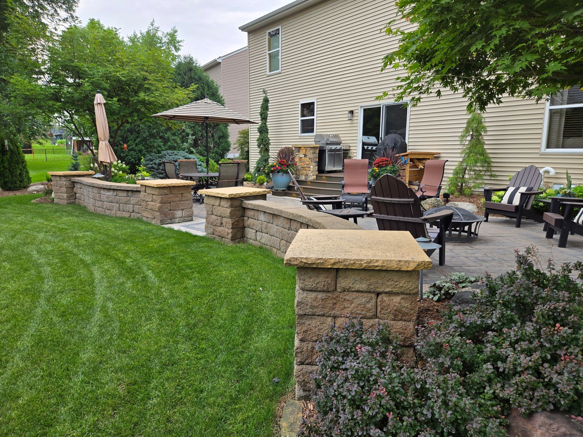 Backyard patio with stone walls, seating, and landscaping; house in background.