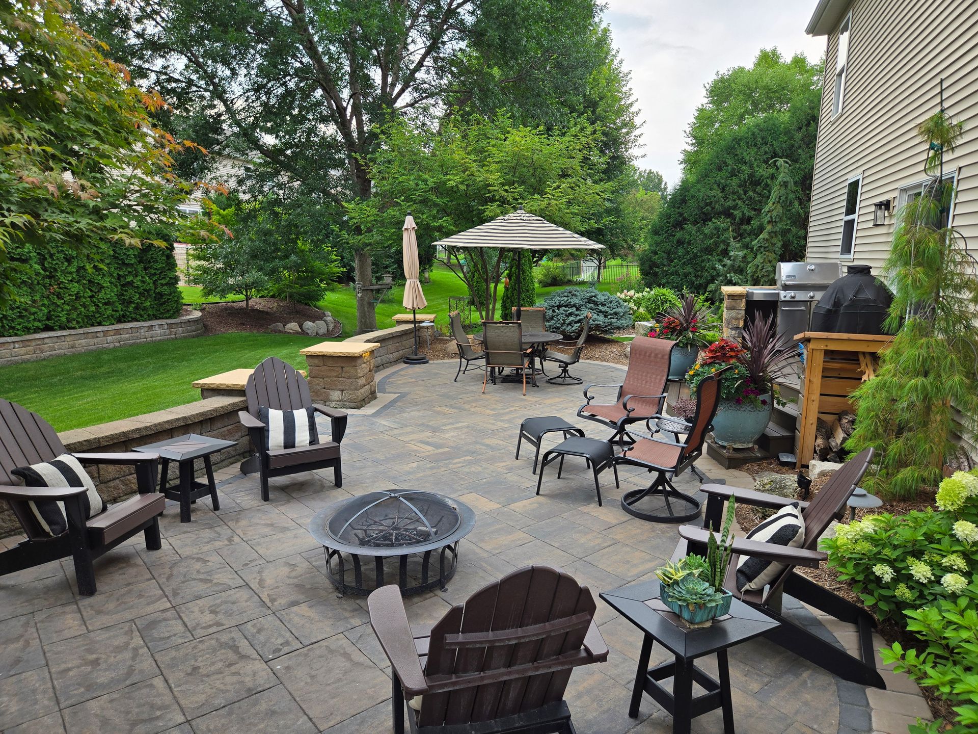 Patio with Adirondack chairs, fire pit, and umbrella table in a backyard with lush greenery.