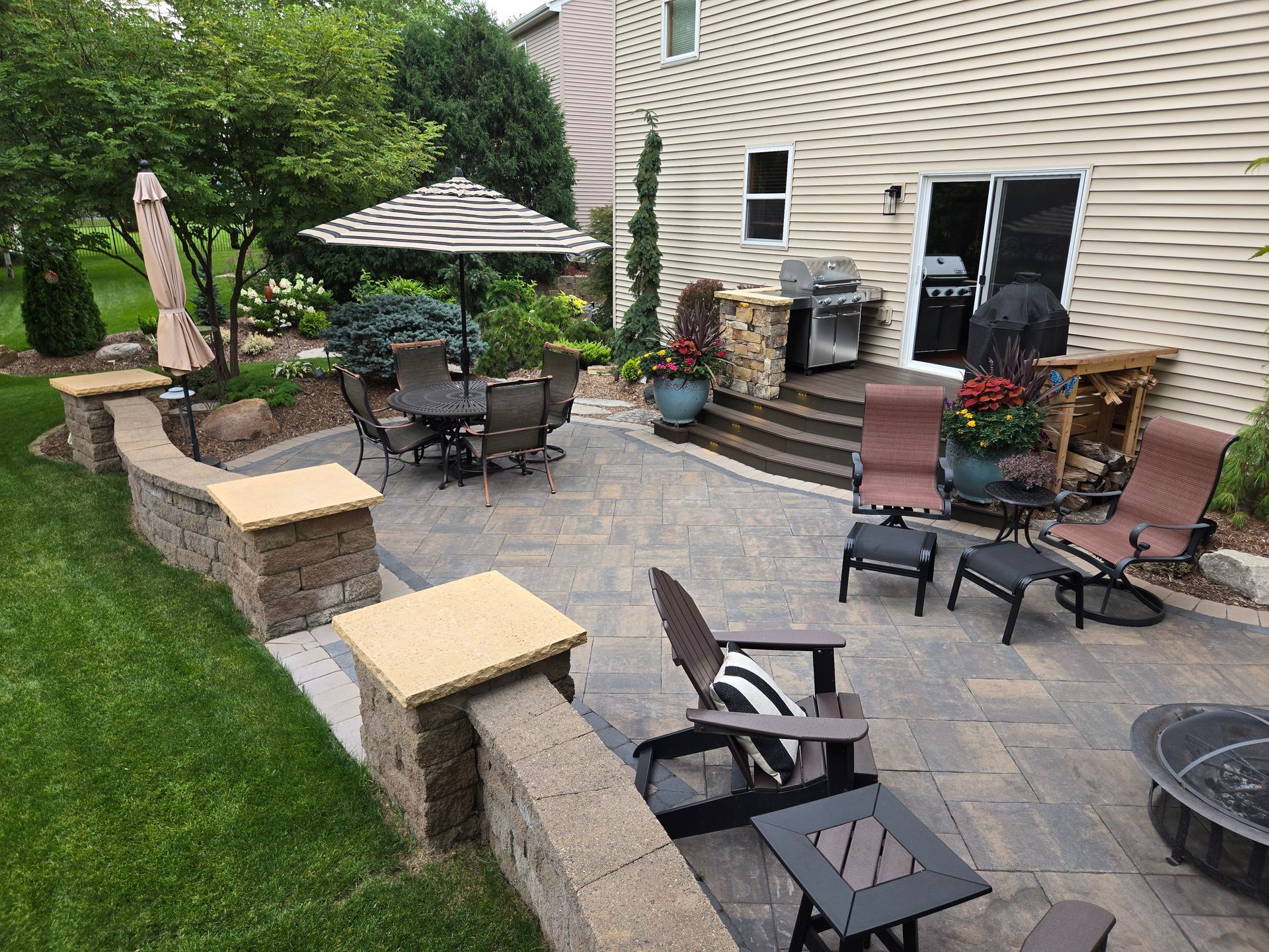 Patio with seating, grill, and fire pit. Brown pavers, retaining wall, and green lawn.