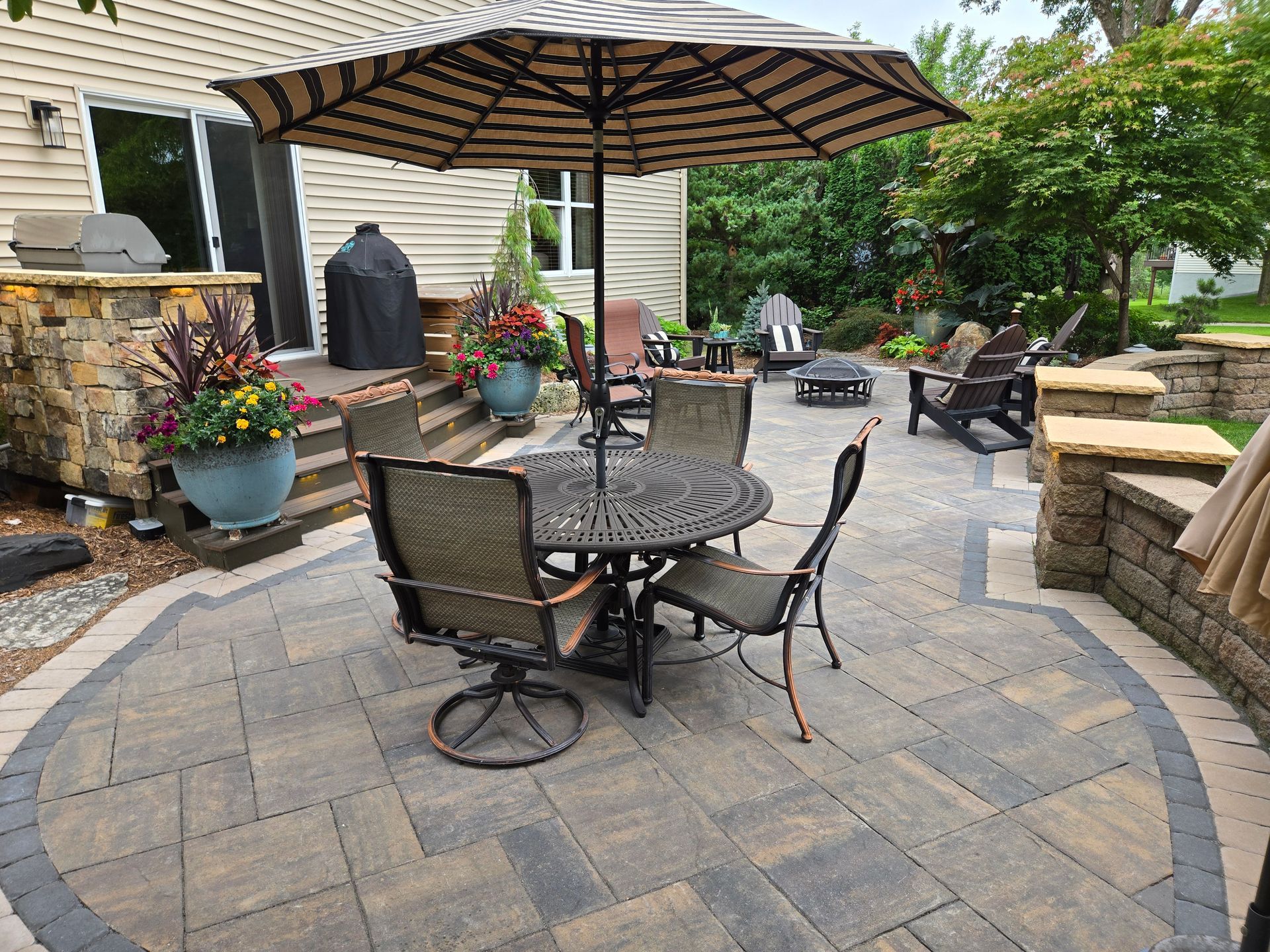 Patio with outdoor dining set under umbrella, stone walls, and surrounding landscaping.