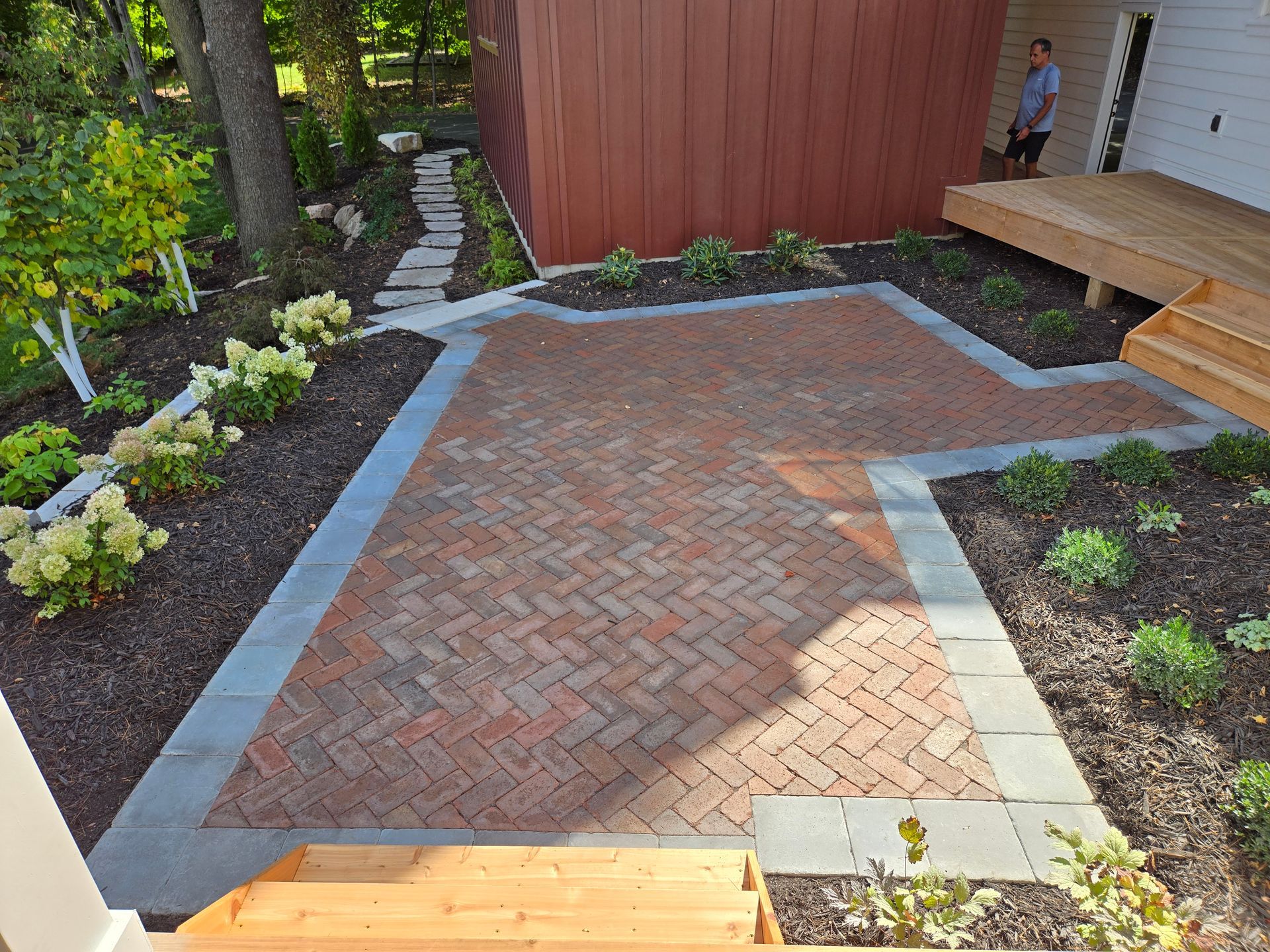 Brick patio with herringbone pattern, edged by gray concrete, adjacent to a wooden deck and building, with landscaping.