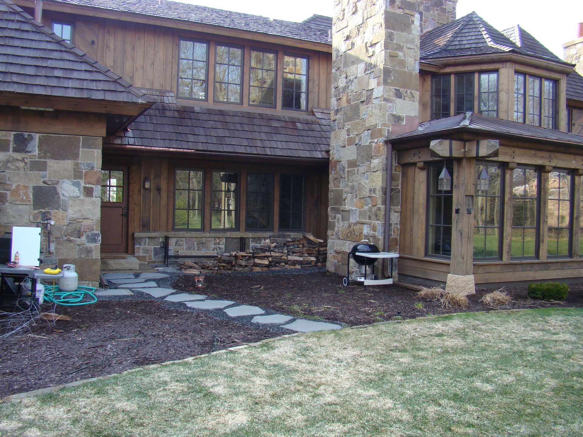 Brown wooden house with stone chimney and walkway, firewood, and grill on the lawn.