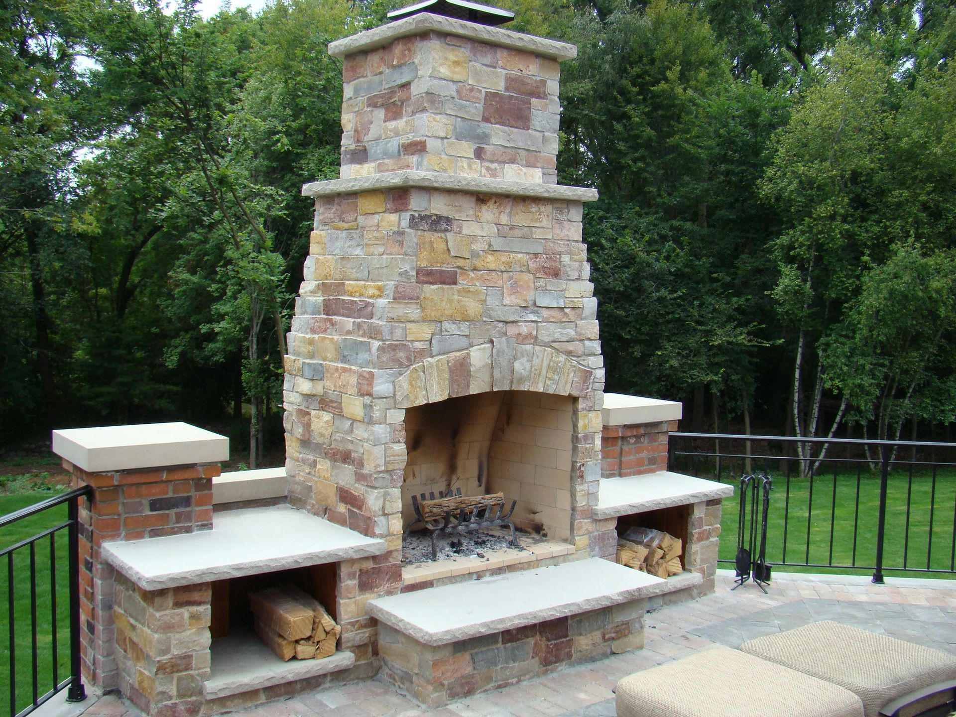 Stone outdoor fireplace with wood storage and chimney, set on a patio with a grassy background.