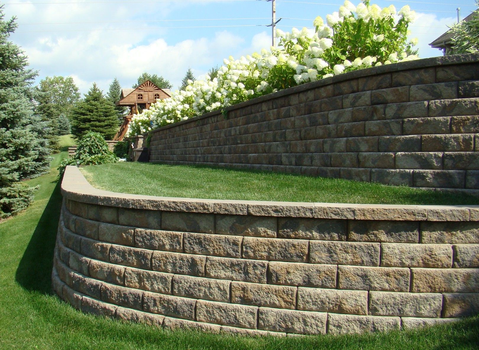 Curved retaining wall of beige blocks with green grass and white flowering bushes on top.