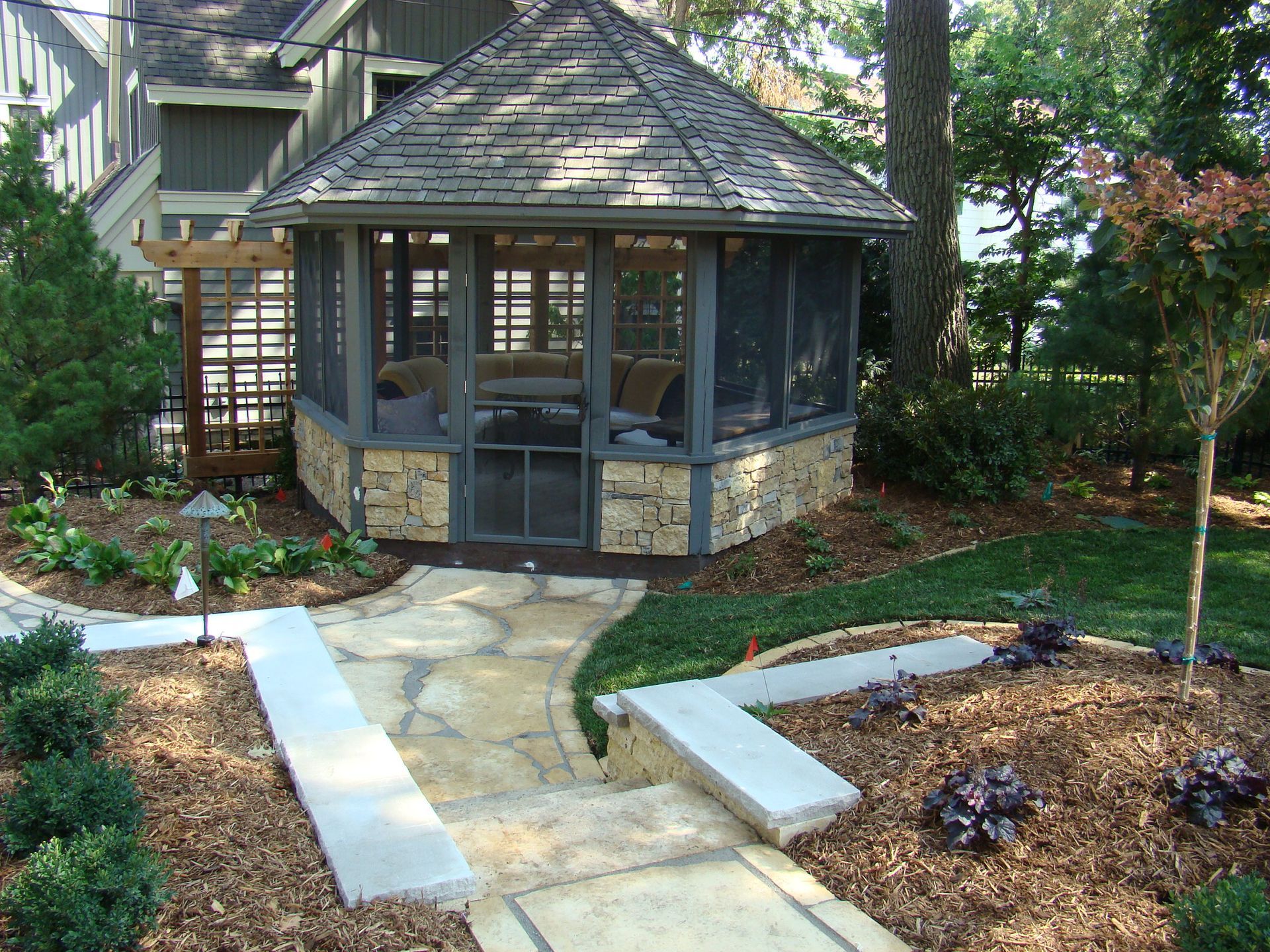 Gazebo with stone base, screened windows, and shingled roof, set in a landscaped yard with a stone path.